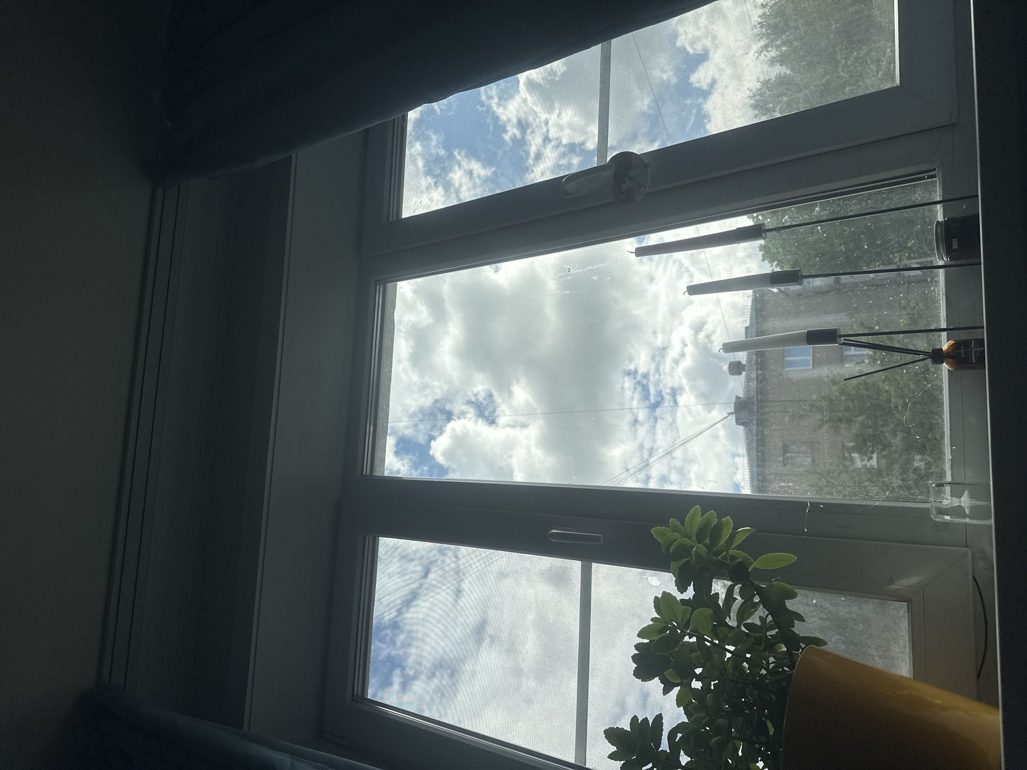 This is an interior, close-up shot of a modern window with a view of the sky and distant buildings, taken from inside a room in Khimki, Russia. The window is divided into multiple panes, with a clear view of a bright, cloudy sky. Sunlight streams through, illuminating the clouds and creating a somewhat hazy effect.
The subjects within the frame include the window itself, which appears to be made of white PVC, and its hardware, such as a white handle. Hanging from thin wires across the lower panes of the window are several reed diffuser sticks, some with what appear to be white reeds and others with darker tips. There are also a couple of glass jars visible. On the right side of the lower pane, a potted plant with vibrant green leaves is placed in a bright yellow pot. The plant is partially obscuring the view of the outside.
The scene suggests daytime, likely morning or afternoon, given the bright sunlight and clouds. The weather appears to be partly cloudy. The presence of buildings and trees in the background provides a glimpse of an urban or suburban environment. The overall mood is calm and domestic, with a focus on the interplay of light and shadow. The image is oriented vertically. There is no visible text in the image.