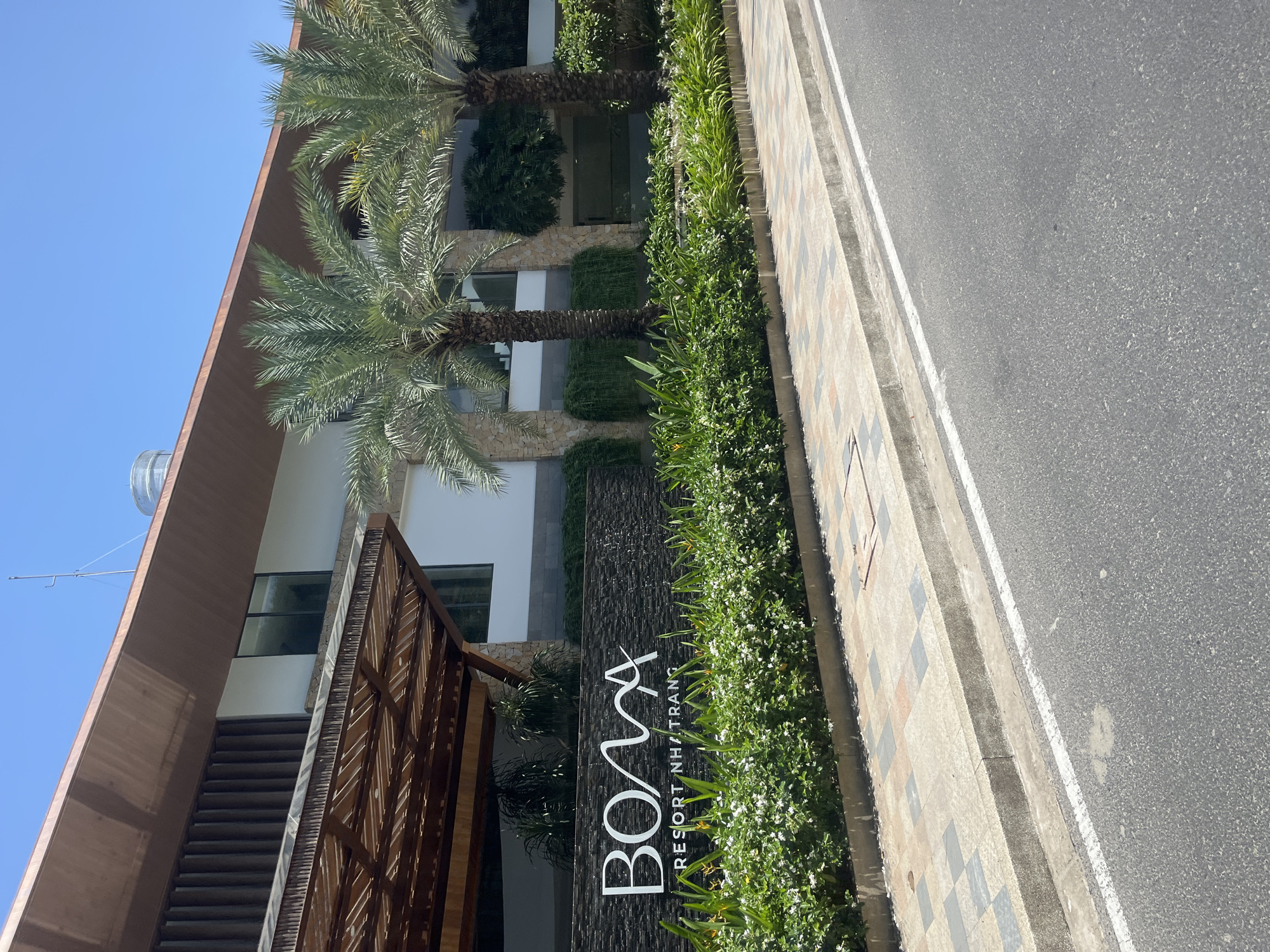 The image shows the entrance to the BOMA RESORT NHATRANG in Nha Trang, Vietnam. The resort's name is prominently displayed in large white letters on a dark, textured wall, below which is a shorter line of text. The entrance is flanked by lush green bushes with small white flowers and tall palm trees. Behind the landscaping, the modern architecture of the resort is visible, featuring stone accents, large windows, and a wooden-covered entryway. The scene is bathed in sunlight under a clear blue sky, with a paved sidewalk and an asphalt road in the foreground. No people are visible in the image.
