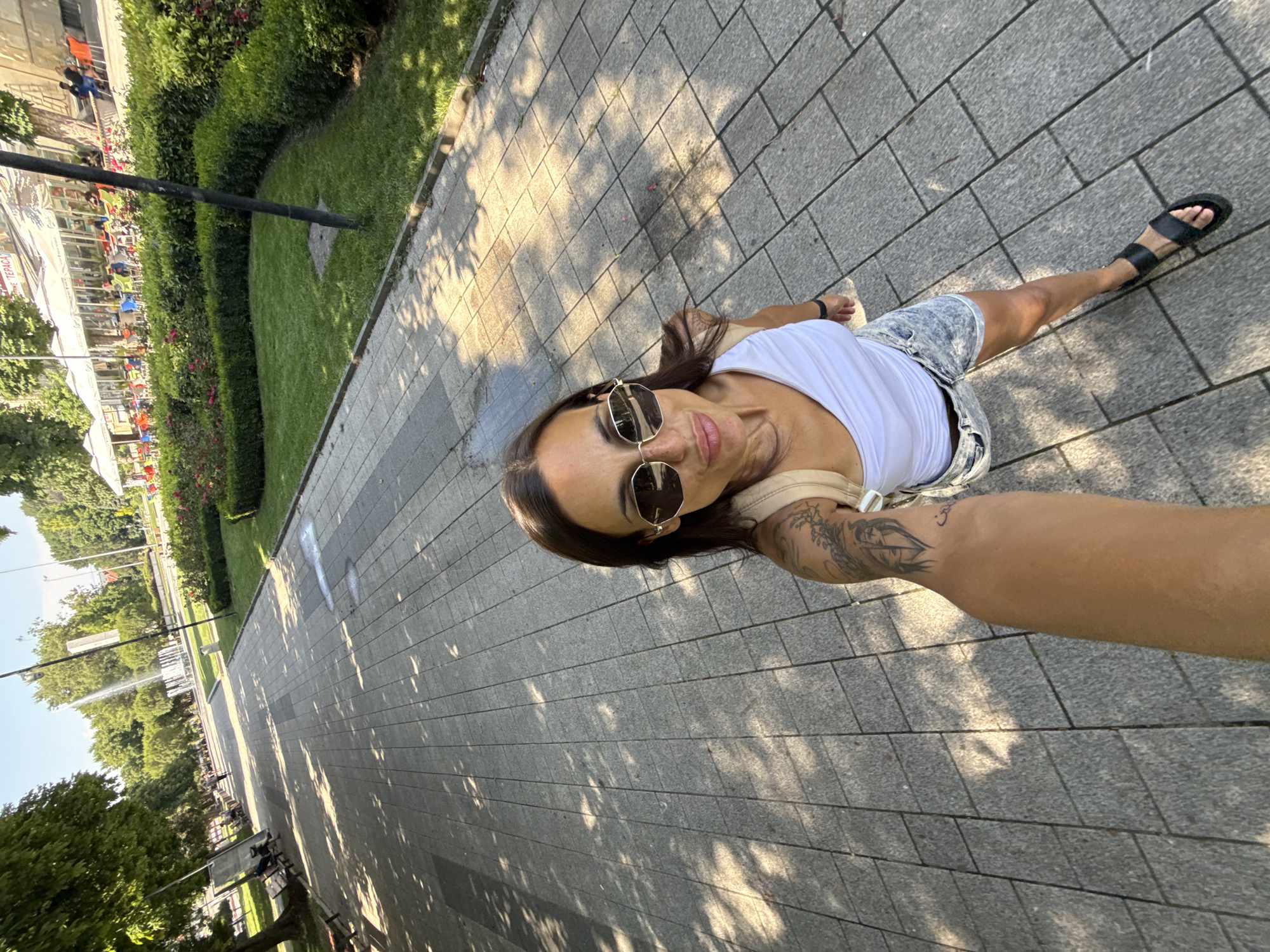 In Pleven, Bulgaria, a woman with sunglasses and a white top takes a selfie in a park. She is wearing denim shorts and black sandals. The park features a paved walkway, manicured bushes, a fountain, and trees. In the background, there are cafes with outdoor seating and people visible. The sun casts shadows on the walkway.