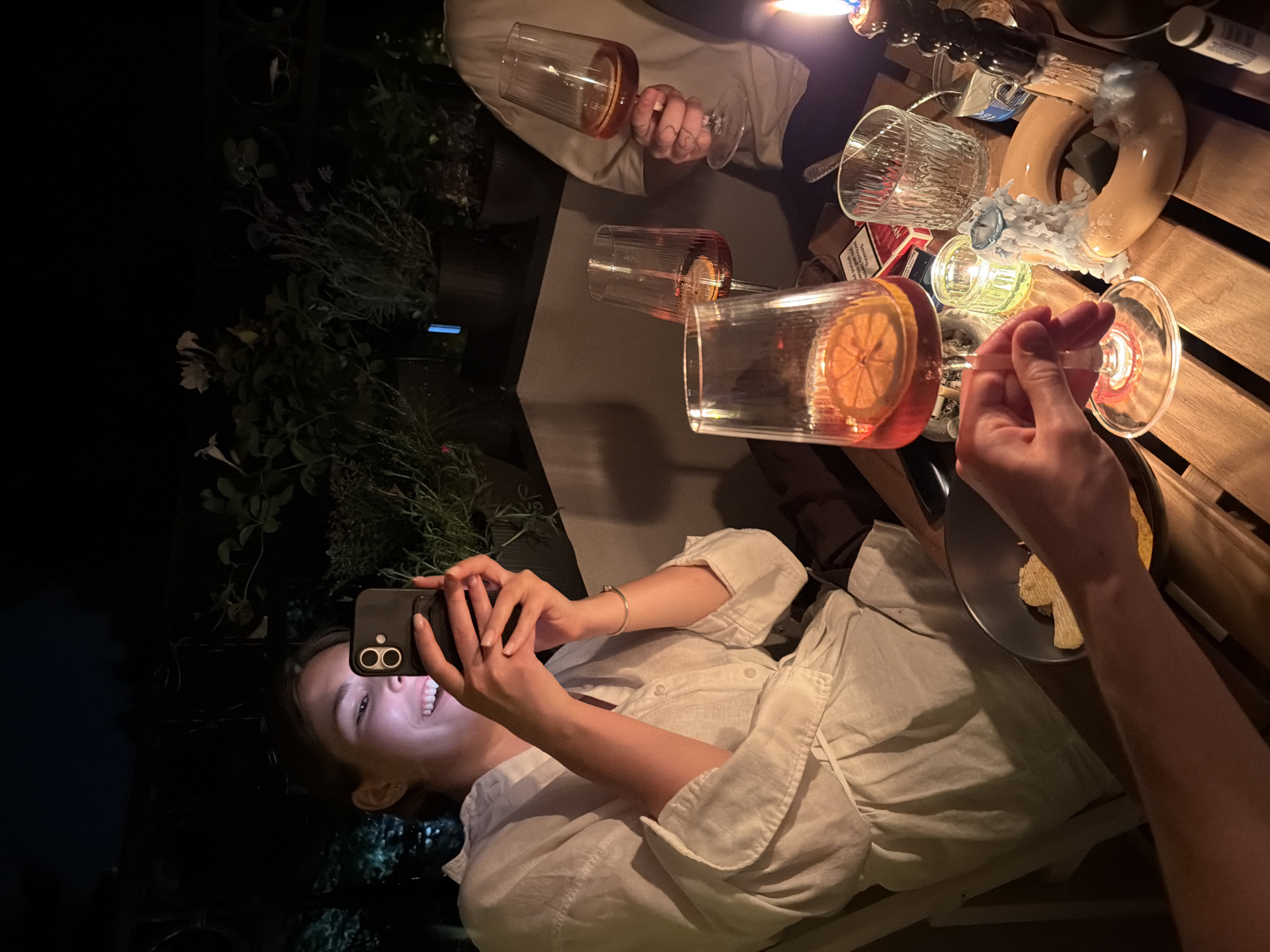 At night, a person is taking a picture with a smartphone of a gathering around a wooden table laden with glasses of drinks, candles, and food. The location is likely a patio or outdoor area in Palilula Urban Municipality, Serbia, evidenced by the presence of outdoor plants and the relaxed, social atmosphere. The person taking the photo is smiling and wearing a light-colored shirt. Other individuals are present, holding glasses and interacting with the objects on the table. The lighting is dim, provided by candles and the phone's screen, creating a warm glow.