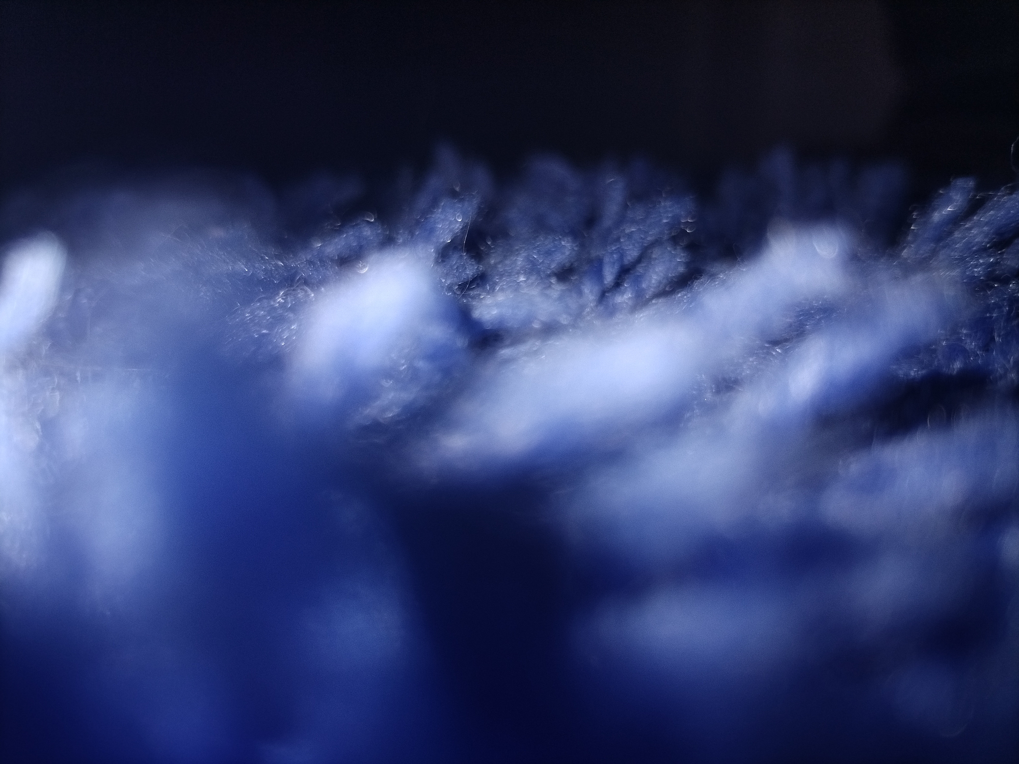 An abstract, macro photograph captures a close-up of textured blue and black material. The lighting creates a blurred effect, with out-of-focus highlights and shadows. The texture appears fibrous or fuzzy, with small, reflective particles or droplets scattered across the surface. The image lacks any discernible location or context, as it is abstract and devoid of identifiable objects or features.