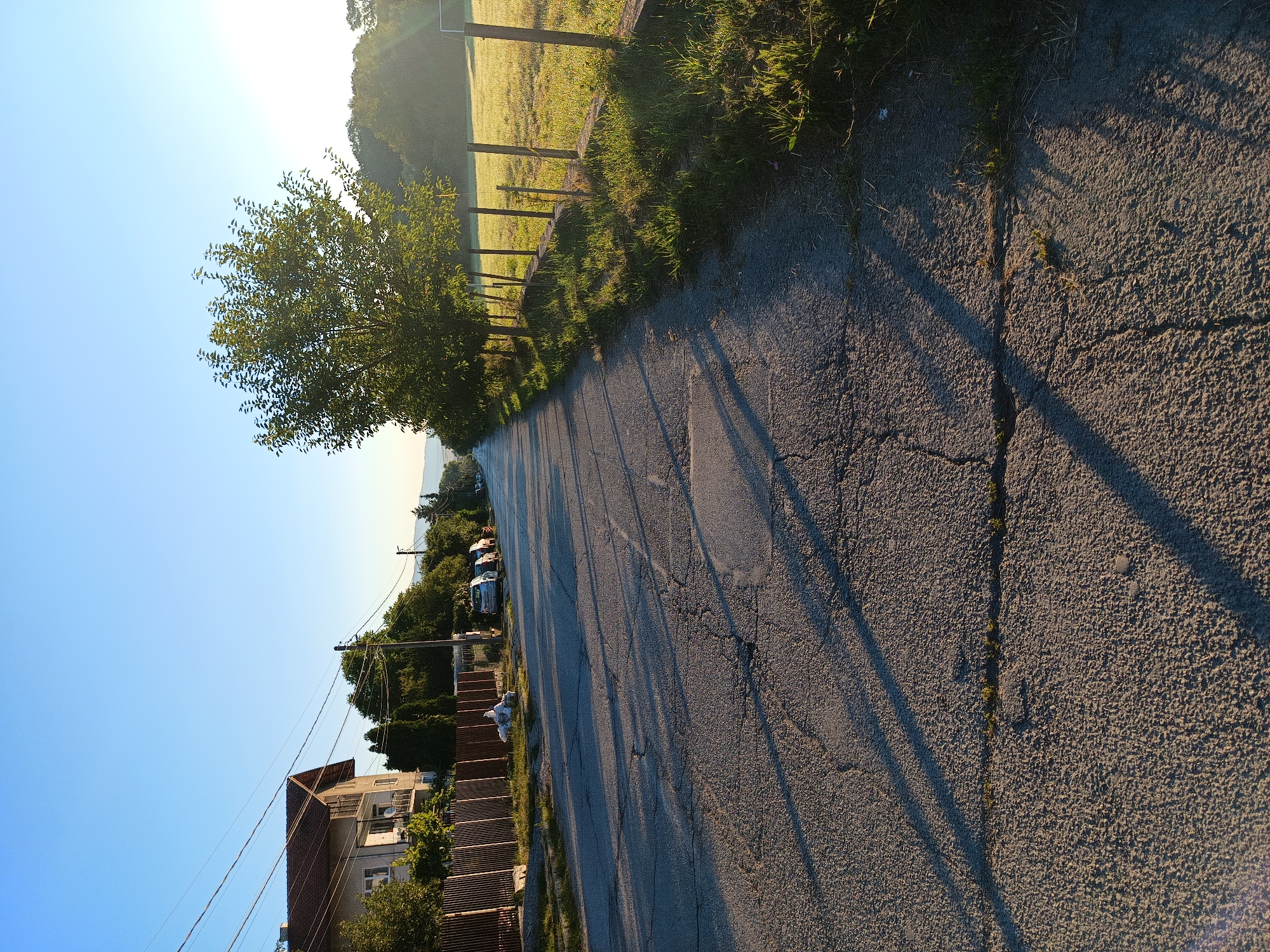 The image shows an asphalt road in Kralev Dol, Bulgaria, on a clear day with a bright blue sky. The road is lined with trees on the right and a residential property with a fence and a house on the left. Several cars are parked along the left side of the road. The sun is casting long shadows across the road, indicating it is likely early morning or late afternoon. The environment appears to be rural or semi-rural, with a grassy field visible to the right of the road. The road surface shows signs of wear and tear, with visible cracks and patches.
