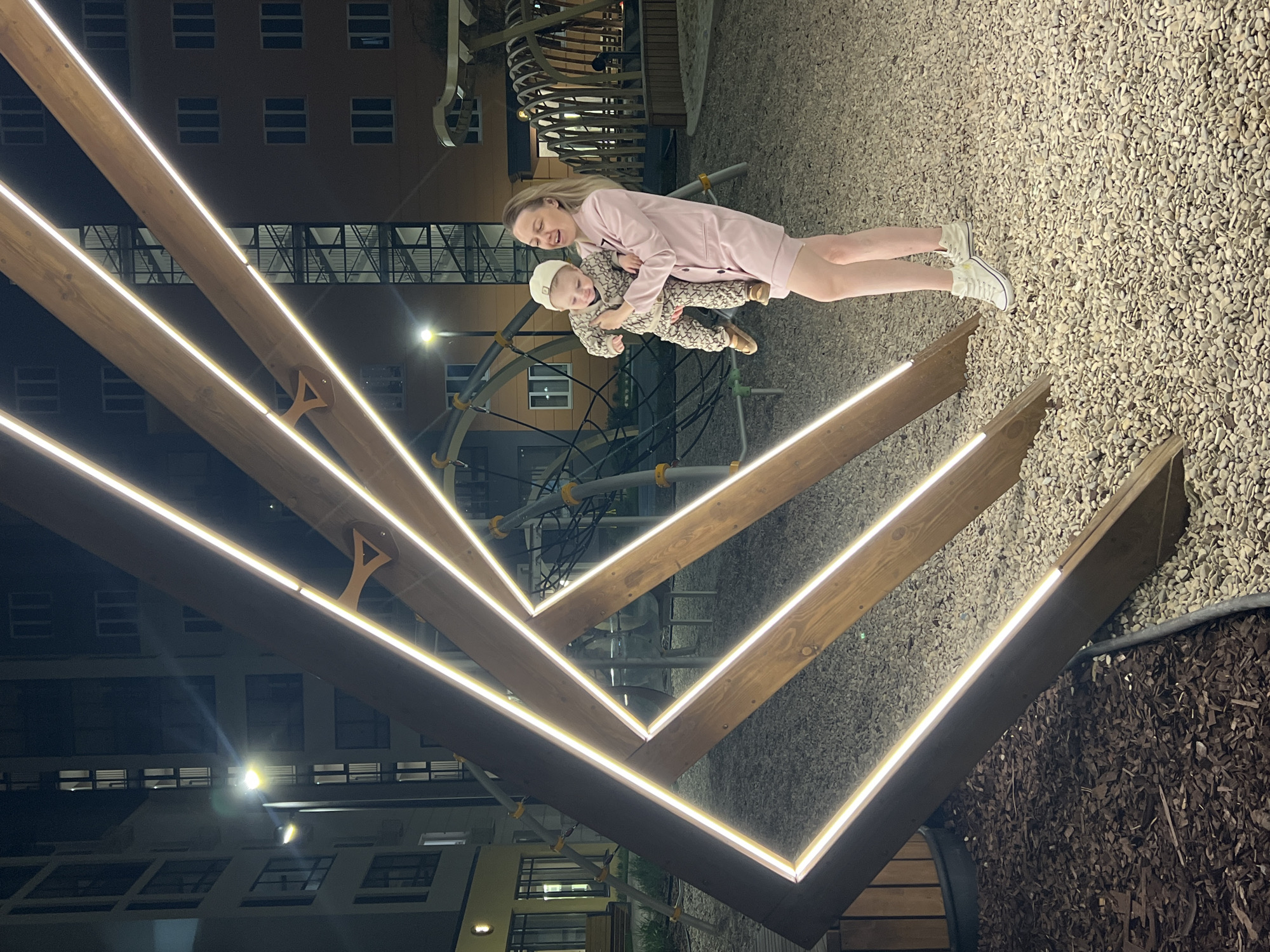 In the city of Pervomaysky, Russia, a mother wearing a light pink dress holds a baby dressed in a patterned outfit and a white hat. They stand on a playground covered in gravel. Behind them is a climbing structure and a building with lit windows. A geometric structure made of wooden beams and LED lights is in the foreground.