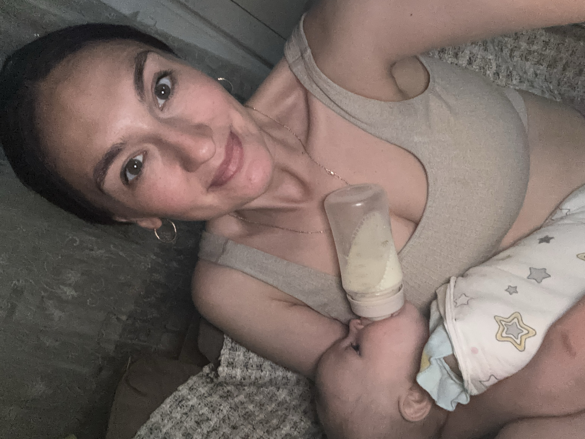 A woman is lying down and bottle-feeding a baby. The woman has dark hair pulled back, and she is wearing a beige sports bra and a gold necklace with a small pendant. She is smiling and looking at the camera. The baby is wrapped in a blanket with stars on it and is drinking from a bottle. The background is dark and blurry, but appears to be a textured wall. The location is not discernible as Sochi, Russia, from the available image data.