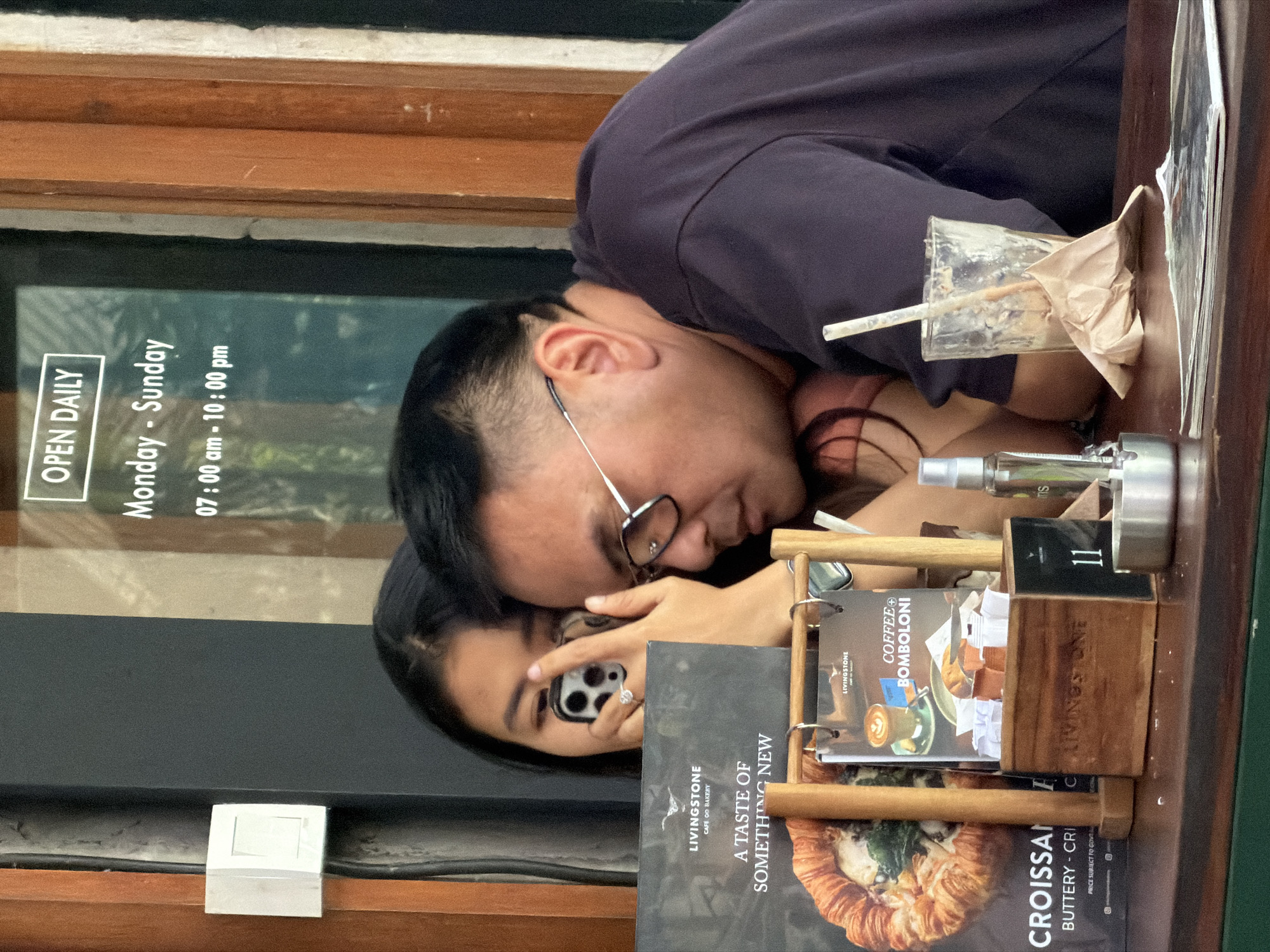 In North Kuta, Indonesia, a man and a woman are seated at a table, with the woman holding up a smartphone to take a photo. The man, wearing glasses and a dark t-shirt, leans his head towards the woman. In front of them are menus, a partially consumed beverage in a glass with a straw, and an ashtray. Behind them, a window displays the text "OPEN DAILY" and "Monday - Sunday 07:00 am - 10:00 pm". The environment appears to be an indoor seating area, likely a cafe or restaurant.