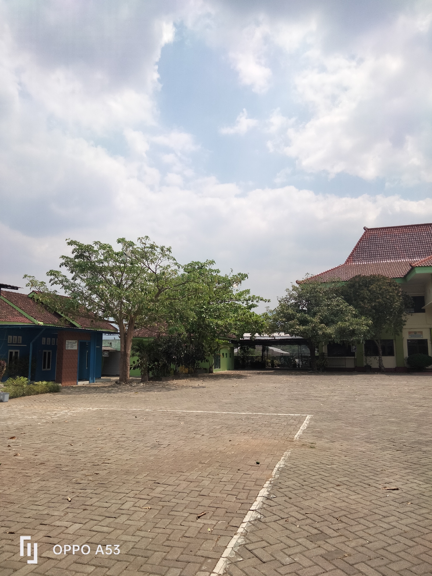 The image depicts an outdoor scene in the city of Karangan, Indonesia. The foreground is a paved courtyard area made of interlocking brick pavers, with some scattered leaves. White lines mark out a section of the paved area. In the middle ground, there are several buildings and trees. To the left is a blue building with a red-tiled roof, featuring several windows. Next to it is a larger tree with green foliage. Further to the right, there is another building with a red-tiled roof and green accents. A cluster of trees with green leaves is situated in front of this building. The background consists of a bright sky with scattered white and grey clouds. The overall impression is a schoolyard or similar institutional outdoor space. The bottom left corner of the image displays the text "OPPO A53".