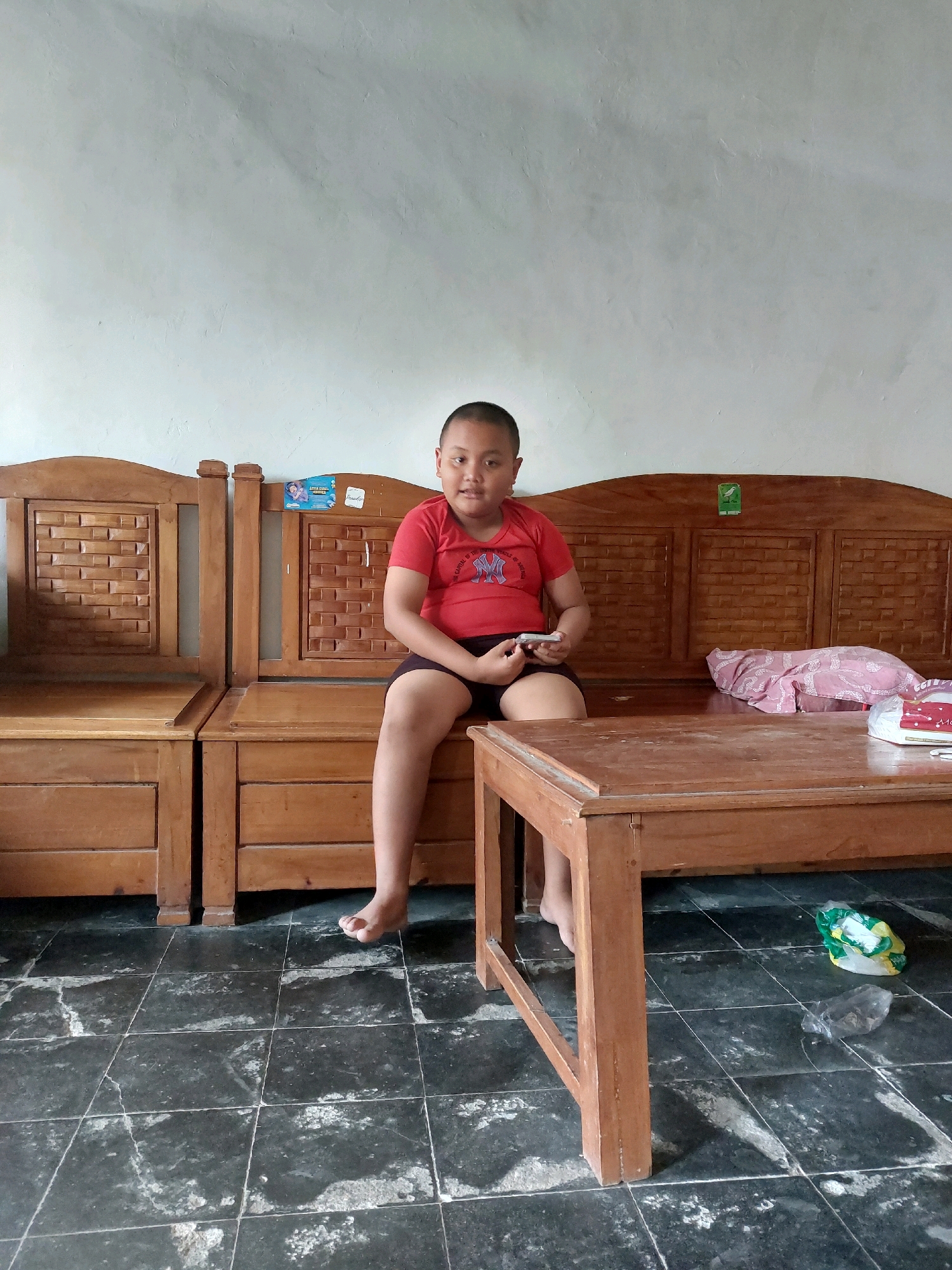 A young person wearing a red t-shirt and dark shorts sits on a wooden bench in what appears to be a living room. The person is holding a mobile phone in their right hand and looking towards the camera with a neutral expression. The bench is made of dark wood with intricate carvings on the backrest and armrests. A wooden coffee table is positioned in front of the bench. The floor is tiled with dark grey and white squares. In the background, a light-colored wall with a textured finish is visible. There are some items on the bench, including a pink cloth and some white bags. On the floor near the coffee table, there are two plastic bags. The overall scene suggests a casual indoor setting in a home. The location is identified as SARANGAN, Indonesia.