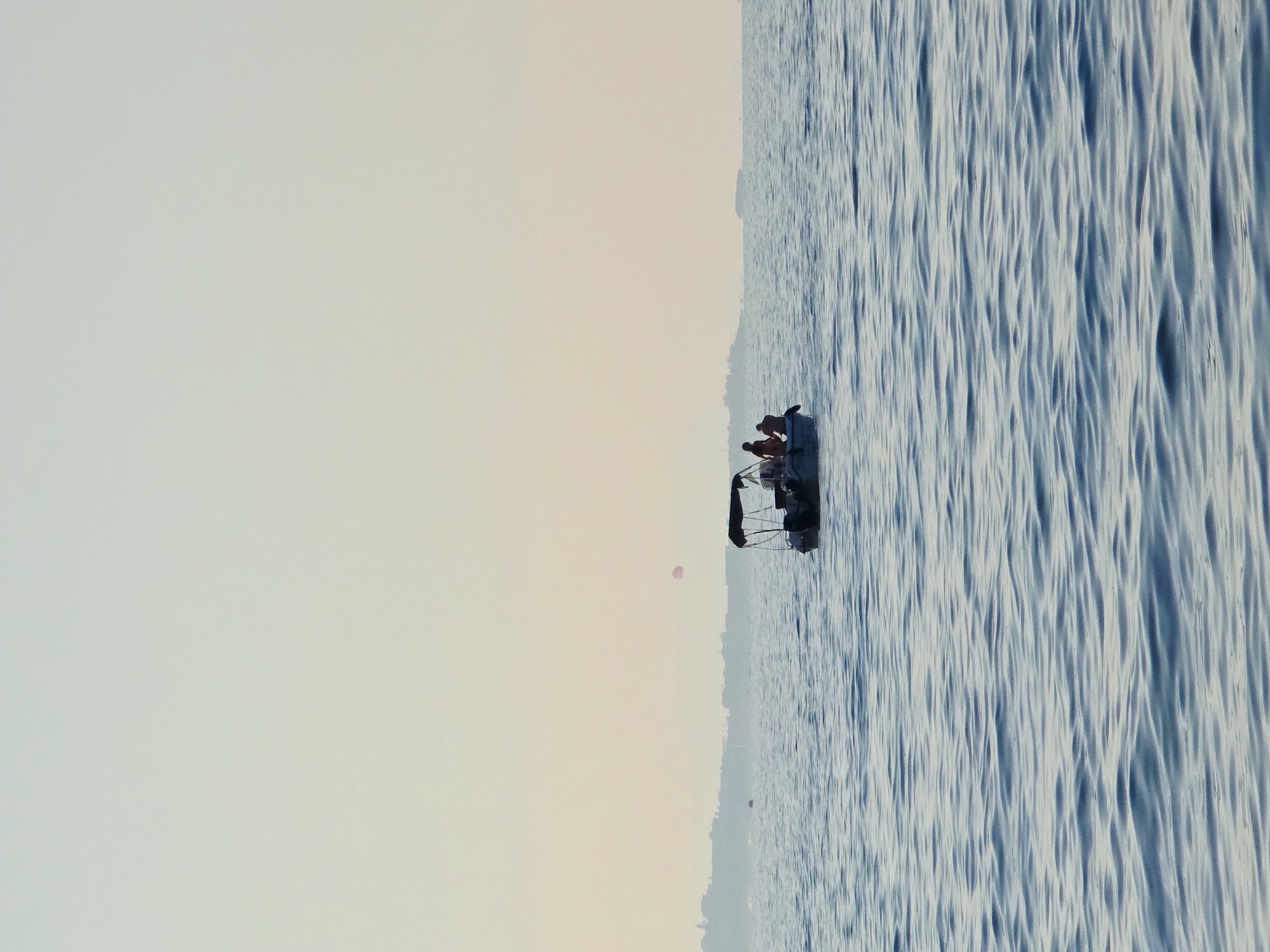 A small motorboat with a canopy is on the water, with several people visible in it. The boat is in the middle distance, with choppy blue water in the foreground and a hazy coastline with some structures in the background. A small pink parachute is visible in the sky above the coastline. The sky is a pale yellow-white, suggesting it is either sunrise or sunset. The scene appears to be in or near the city of Malia, Municipal Unit, Greece, given the coastline and the general ambiance.