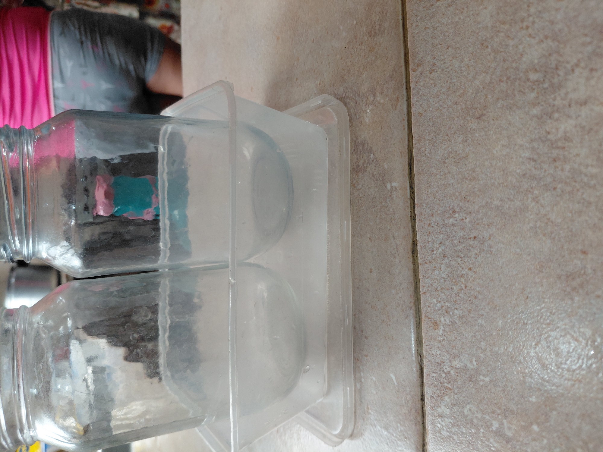 The image, oriented sideways, displays two clear, empty glass jars lying horizontally within a transparent plastic container. Each jar features a screw-top neck. The plastic container is rectangular with slightly rounded corners, and the jars rest in molded depressions within it.
In the blurred background, a person is partially visible from the back, wearing a pink top over a grey garment. The rest of the background is indistinct, suggesting an indoor setting.
The objects are positioned on a light beige or off-white tiled surface, which exhibits subtle variations in color and texture, consistent with ceramic or porcelain tiles. A dark grout line is visible vertically along the right side of the tiled surface.
No specific activity is occurring; the scene is static. Faint, embossed text, possibly indicating product details such as "PHTHALATE FREE," can be minimally discerned on the side of the plastic container. The overall impression is that of a domestic indoor environment, potentially a kitchen or utility area, in Santa Ana, Venezuela.