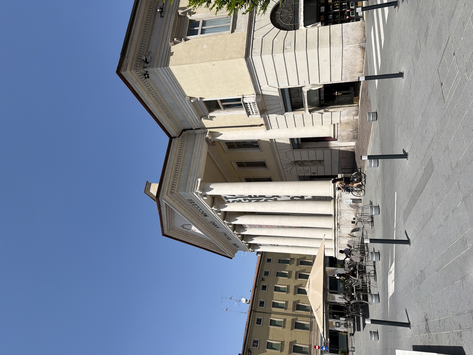 The image is a low-angle, upward-facing shot of a neoclassical building with prominent columns, located in Mantua, Italy. The sky is clear and blue. The building features a triangular pediment adorned with inscriptions and details, supported by multiple white columns. The architectural style appears to be Italian.
To the right of the neoclassical building is a multi-story building with a more utilitarian design. Below and in front of the buildings is a public plaza area with gray pavement. The plaza includes metal posts and outdoor cafe-style seating (tables and chairs). There are a few people visible in the scene. One person is riding a bicycle and several others are seated at the outdoor tables. A portion of a beige sunshade is visible. A pedestrian crosswalk is also visible.