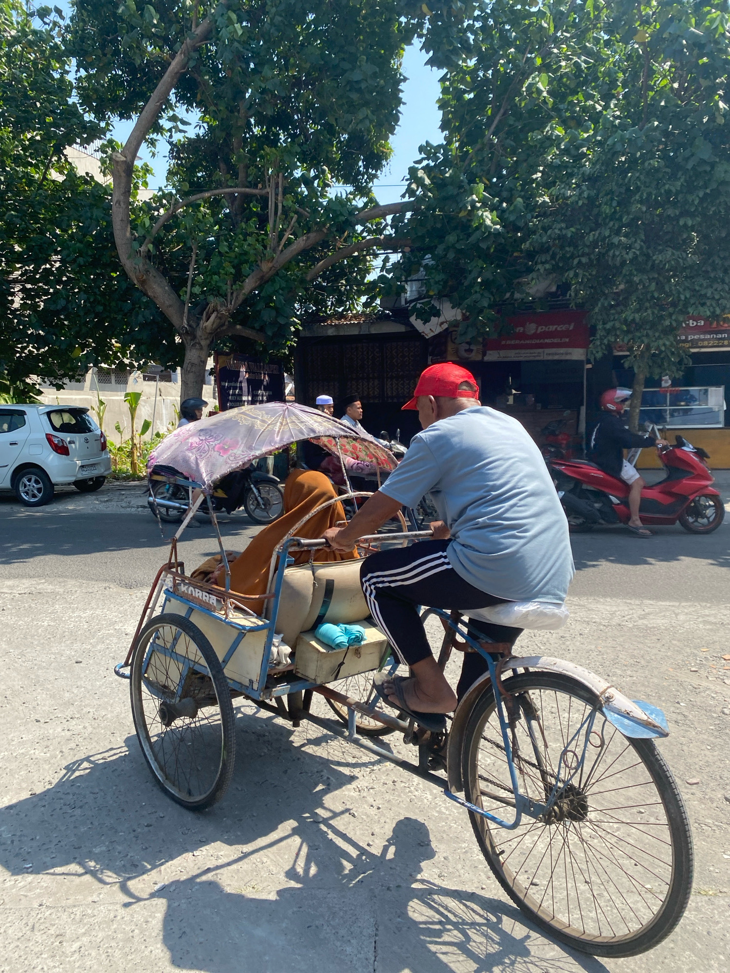 In Kediri City, Indonesia, a male pedicab driver wearing a blue shirt, black pants with white stripes, and a red cap pedals a pedicab carrying passengers. The pedicab has a light-colored canopy decorated with pink floral patterns and is adorned with the name "KORRA" on its side. A passenger, covered in a tan-colored garment and headscarf, sits in the pedicab. In the background, a white car is parked on the left, and a red motorcycle is seen on the right, with buildings and lush green trees lining the street. The scene is set on a paved road under a clear blue sky, suggesting a sunny day.