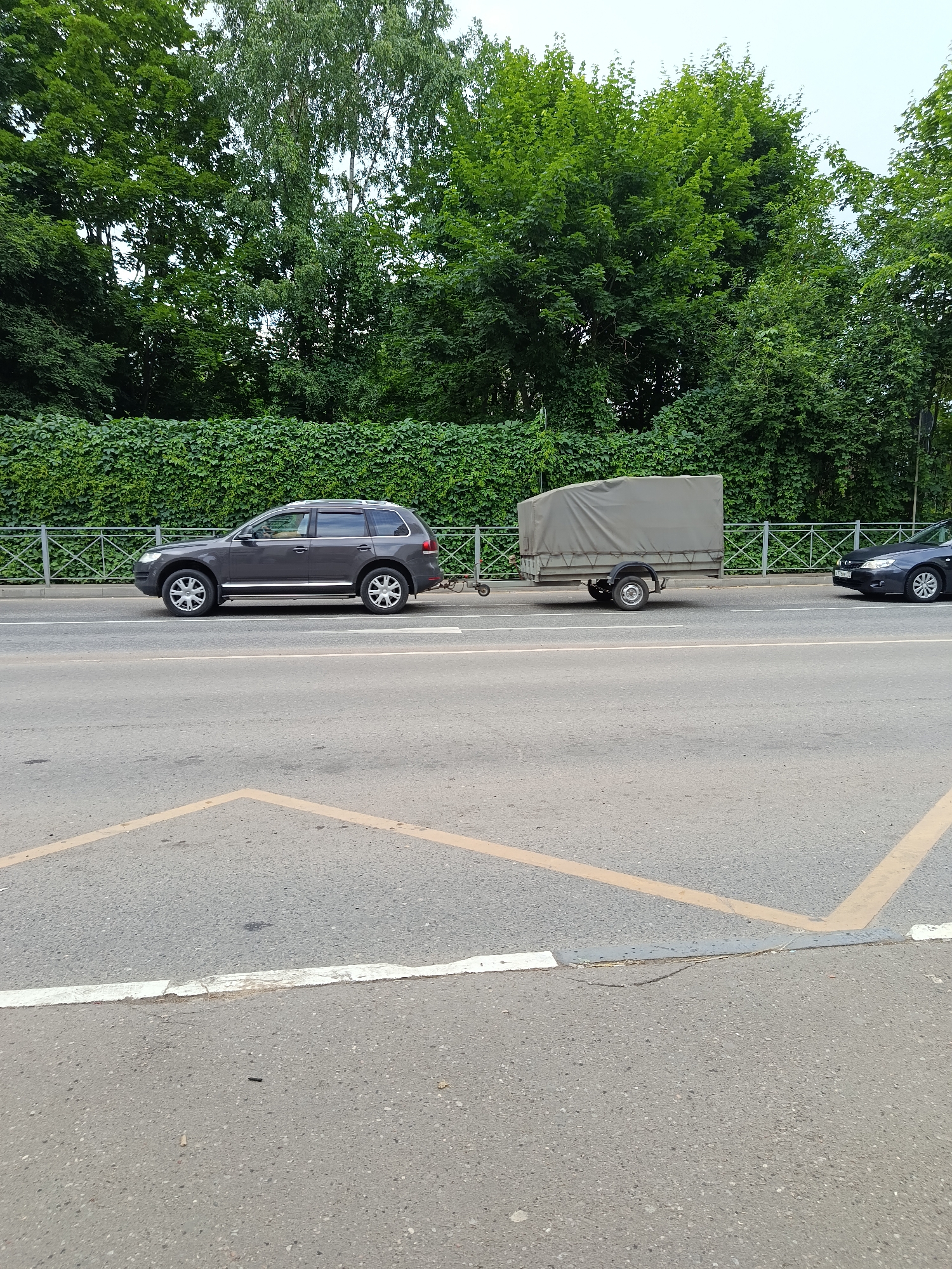 A dark gray SUV is driving on a road, towing a small, light gray trailer. The SUV is in the left lane, and the trailer is attached behind it. The trailer is covered with a tarp. The road appears to be an asphalt surface, and there are yellow and white markings on it, suggesting it's a street or highway. In the background, there is a dense green hedge and a line of trees with lush green foliage. The sky is overcast, indicating a cloudy day. The scene is captured from a side-angle perspective, showing the vehicles from their left sides. The overall impression is of a typical road scene on an overcast day, with vehicles in motion.