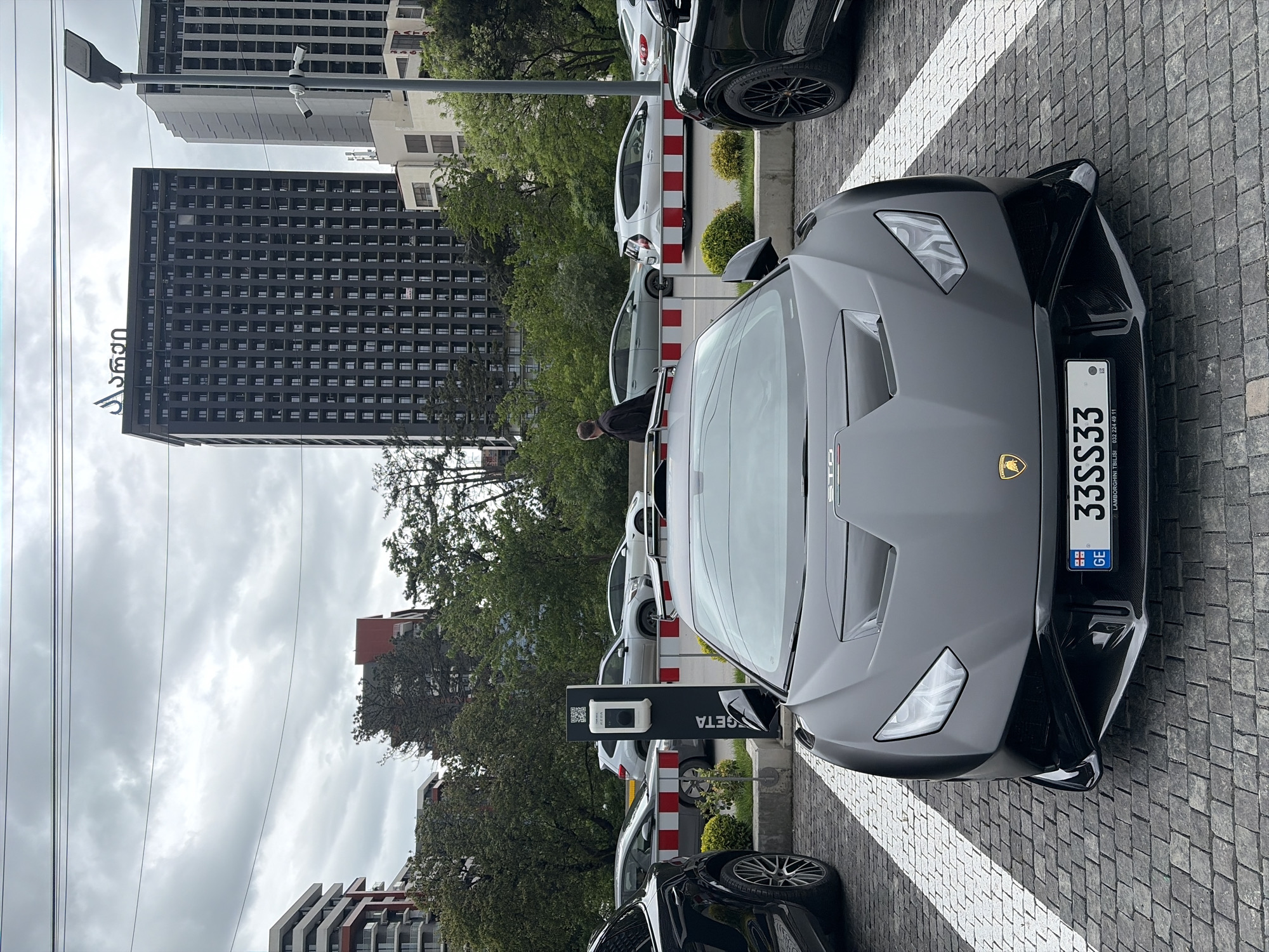 The image shows a gray Lamborghini parked on a brick-paved street, likely in Tbilisi, Georgia. The car has a Georgian license plate that reads "33SS33," and the lower portion of the license plate is labeled "LAMBORGHINI TBILISI 032 224 49 11." The "GETA" charging station is visible behind the car. The background reveals modern buildings, suggesting an urban setting. There are other cars parked around, and trees are present as well. The sky is overcast. The Sofio building is in the background.