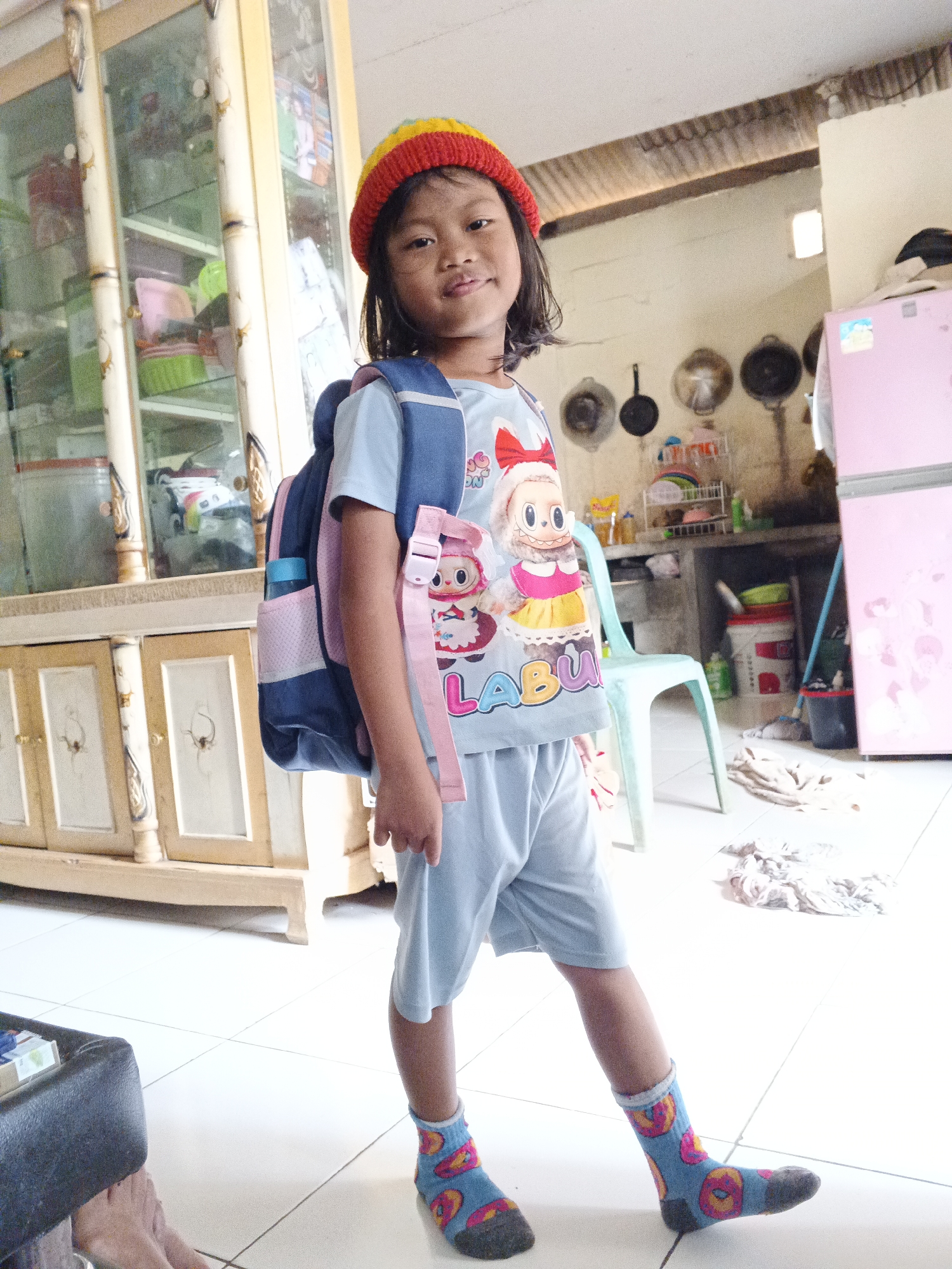 A young girl, appearing to be of Indonesian descent, stands in what appears to be a home environment, possibly in the city of Warungkiara, Indonesia. She is wearing a blue t-shirt with cartoon characters, light blue shorts, a multicolored knit hat (red, yellow, and green), and blue socks with a donut pattern. A blue backpack with pink accents is slung over her shoulders. The girl is facing the camera with a slight smile.
The background reveals a cluttered indoor space. To the left, a tall wooden cabinet with glass doors displays various items, including what appear to be kitchenware or food containers. To the right, a pink refrigerator stands against a wall. In the middle ground, a simple teal plastic chair is positioned near a countertop area with cooking utensils and other miscellaneous objects. The floor is tiled in a white grid pattern. The overall lighting suggests it is daytime, with sunlight entering from the right side of the frame. The scene appears informal and domestic.