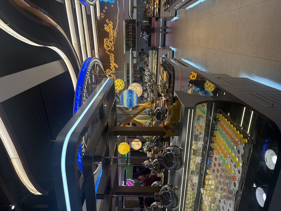 The image shows the interior of a "Royal Buffet" restaurant. There's a contemporary design with curved, illuminated architectural elements. Various food stations are visible, showcasing desserts, salads, and cooked dishes. Some stations have spherical decorations. A "Grillades" sign is also present, and customers can be seen interacting with the buffet offerings.