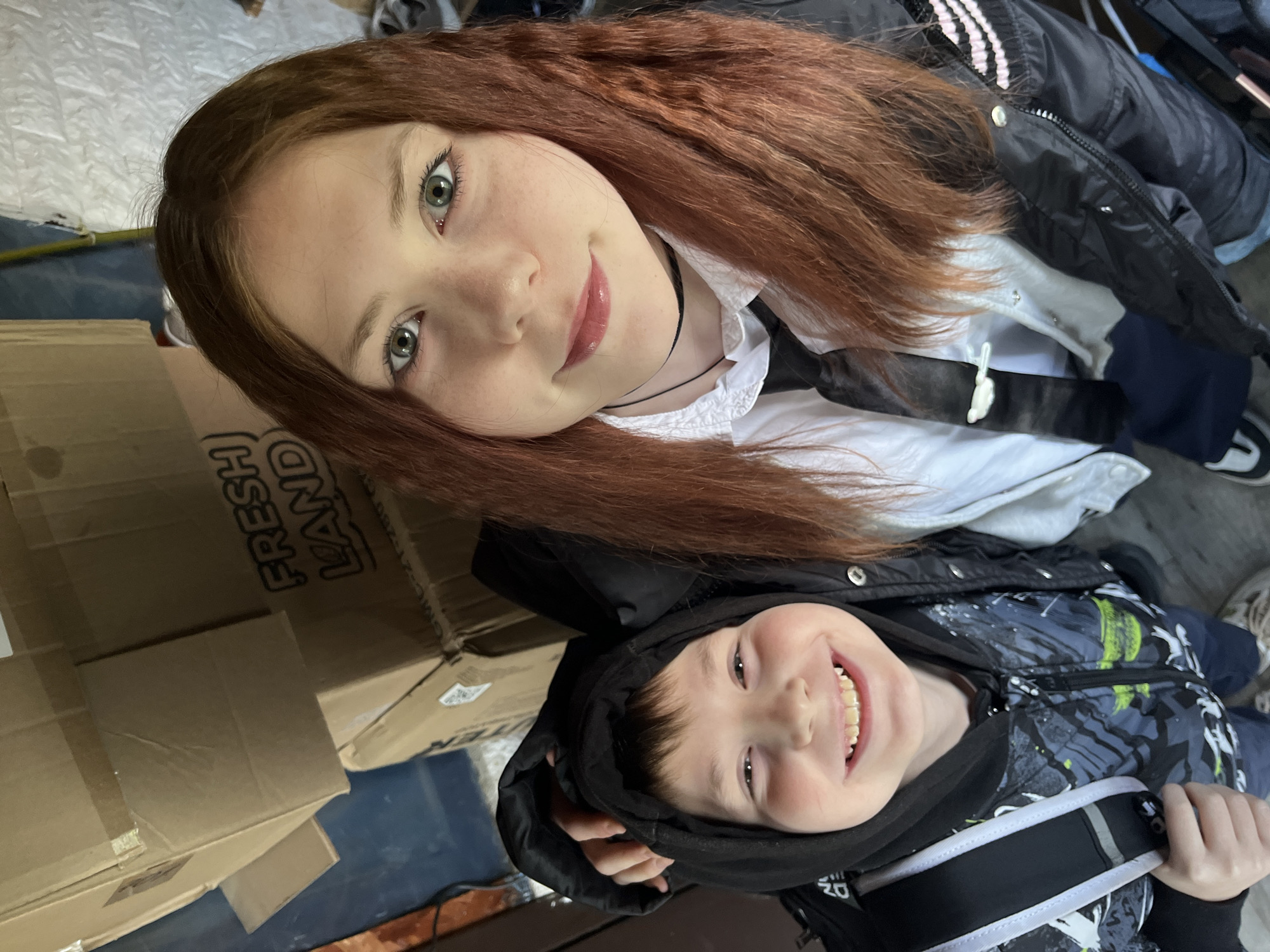 An overhead photograph, rotated 90 degrees clockwise, shows an older girl and a younger boy smiling at the camera. The girl, positioned at the top of the frame, has long, wavy reddish-brown hair and green eyes, with light makeup including pink lip gloss. She wears a white collared shirt, a black choker, and a black jacket. The boy, positioned below her, wears a dark hooded top and a patterned jacket featuring shades of grey, black, white, and lime green, with a visible backpack strap. He has a wide, happy smile, showing his teeth. Both individuals appear cheerful and content.
The background is cluttered, suggesting an indoor or sheltered environment. Several cardboard boxes are visible, with one prominently displaying the text "FRESH LAND". Other parts of the background include what appears to be white plastic sheeting or tarpaulin and dark, indeterminate objects. The lighting suggests an indoor setting, and no specific time of day or weather conditions can be discerned from the image.
