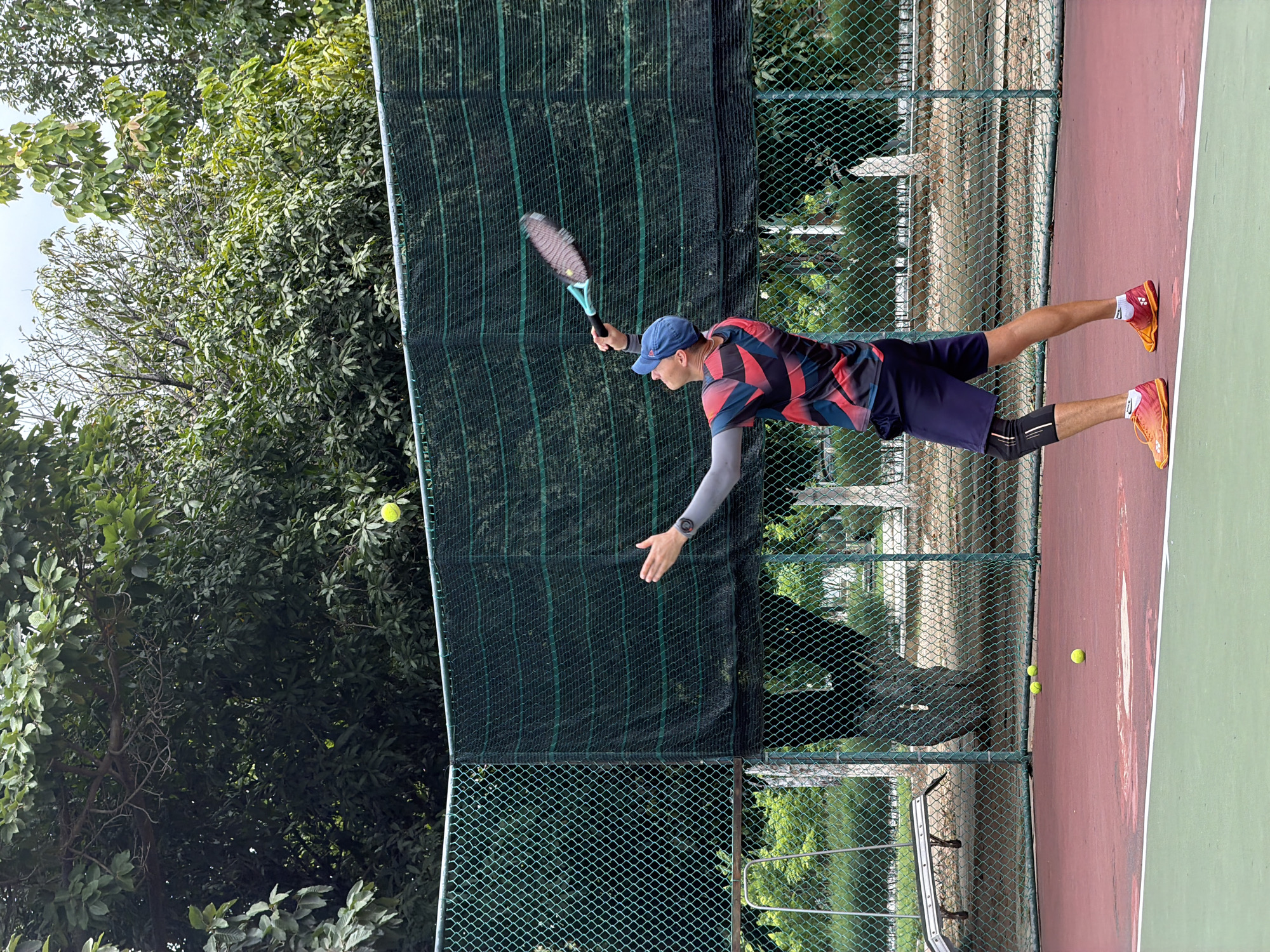 This image captures a male tennis player in dynamic action on an outdoor court, located in Bac Nha Trang Ward, Vietnam. The player is seen mid-swing, leaning forward and extending his right arm with a tennis racket, aiming to strike a yellow tennis ball suspended in the air.
The athlete wears a blue baseball cap with a white Adidas logo, a distinctive red, black, and white patterned athletic shirt, and dark blue shorts. He sports bright orange and yellow Yonex tennis shoes, a grey compression sleeve on his left arm, a black watch on his left wrist, and a black knee brace on his right leg. His expression indicates intense focus on the ball.
The tennis court features a vibrant red playing area in the foreground and mid-ground, transitioning to a lighter green section beyond the baseline. White court lines are visible. A dark green privacy screen, attached to a chain-link fence, forms a barrier behind the player, separating the court from a dense backdrop of lush green trees and foliage, suggesting a natural, park-like environment. Another, less obscured, chain-link fence is visible further back. In addition to the ball the player is targeting, three other yellow tennis balls are scattered on the red court surface in the foreground. The scene is illuminated by natural daylight.