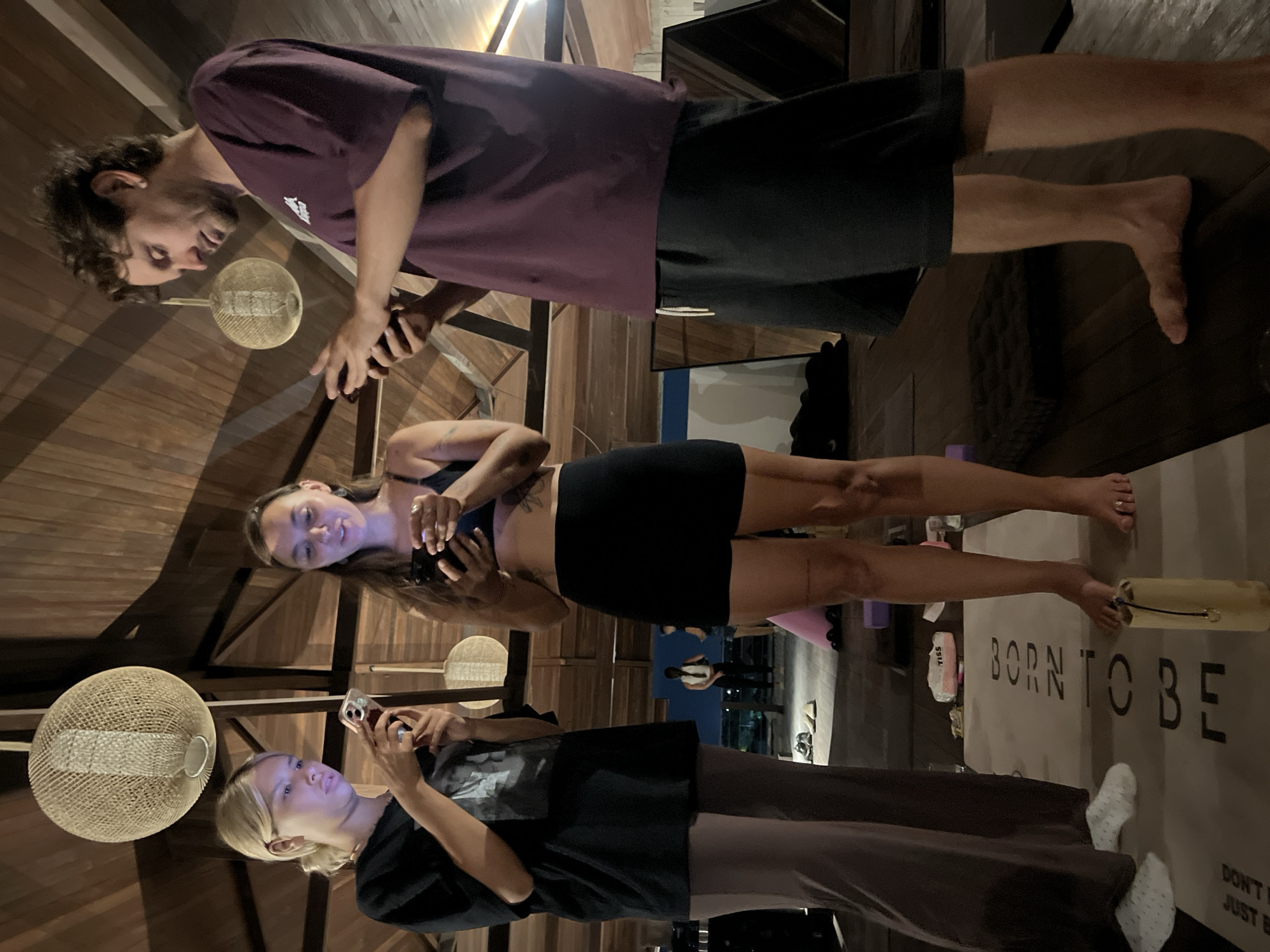 Three individuals are indoors, each engaged with a mobile phone. The setting features a wooden ceiling with pendant lighting. A woman in a sports bra and dark shorts stands barefoot on a yoga mat with the text "BORN TO BE." Another person, wearing dark clothing and pants with white socks, also stands on the mat. A man in a purple shirt and dark shorts stands nearby. The location appears to be in or near the city of Mengwi, Indonesia.