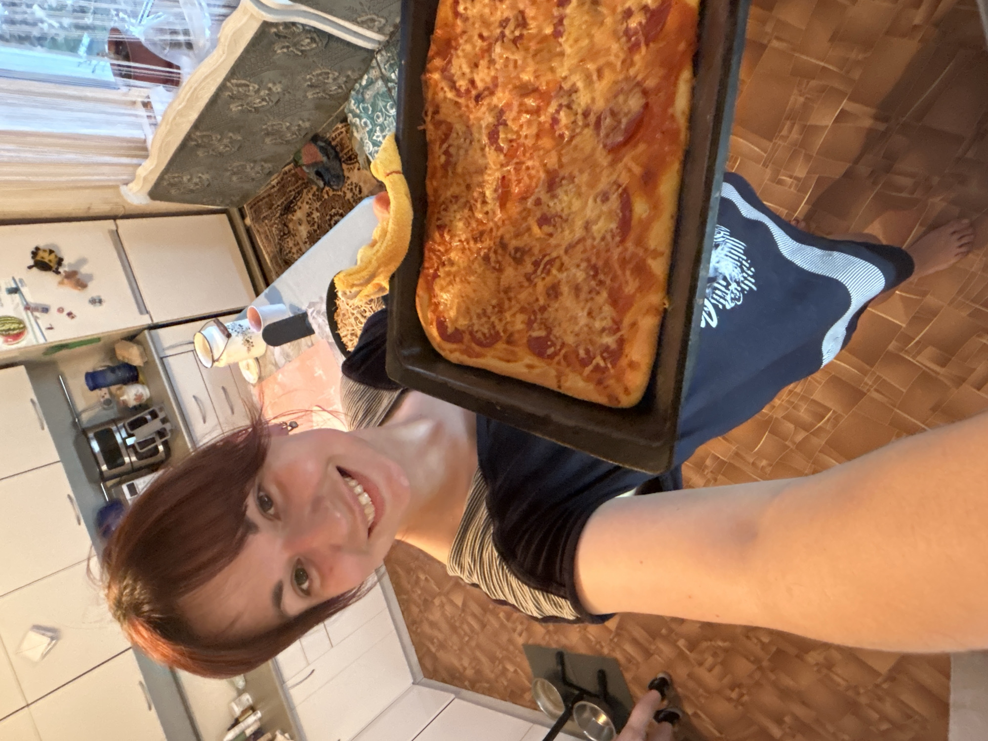 A woman holds a rectangular pizza in a black baking pan. The pizza has a visible layer of melted cheese and what appears to be pepperoni. The woman is standing in a kitchen with white cabinets, and part of a dining area with a table and upholstered chairs is visible in the background. The floor is covered in brown vinyl. The photo is a selfie, taken at an angle looking down, and the woman is smiling. There is no indication of the city Фоки, Russia, in the image.