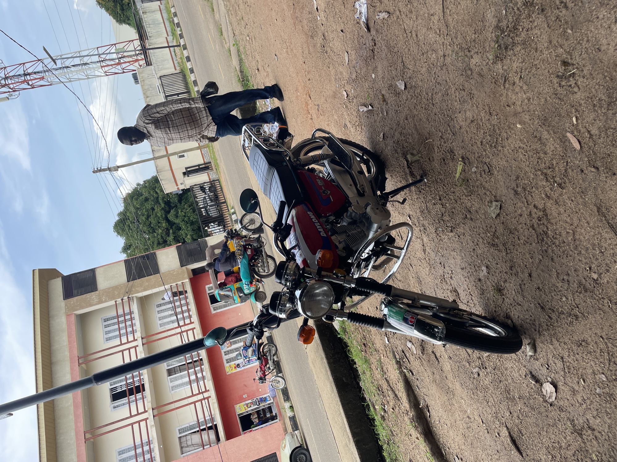 The image depicts a street scene in Jos, Nigeria, focusing on a red motorcycle parked on a dirt shoulder. In the background, a multi-story building with a red and cream facade stands, featuring balconies with white-railed railings. A man wearing a plaid shirt and jeans walks away from the camera towards a cell tower in the distance. A few other motorcycles and a tricycle are also visible on the paved road. The ground is a mix of dirt and sparse vegetation, with scattered leaves and debris. The sky is partly cloudy.