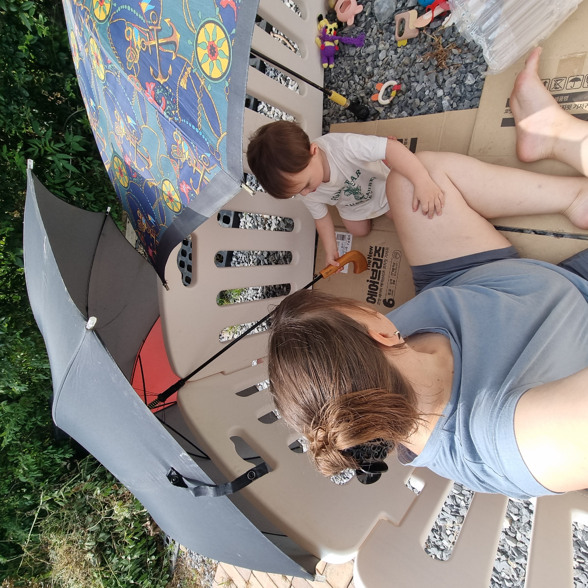 A woman and a toddler are sitting under two umbrellas on a play structure in a yard. The woman is wearing a blue t-shirt and dark shorts. She has her hair pulled back in a bun. The toddler is wearing a white t-shirt and is holding a wooden spoon. The toddler is facing away from the camera. The play structure is made of beige plastic. There are rocks and gravel on the ground around the play structure. The umbrellas are open and providing shade. One umbrella is gray and the other has a nautical pattern. The image is taken from a high angle, looking down at the woman and toddler. The background is blurred green foliage, suggesting an outdoor setting in a yard or garden. The location is Dunnae-myeon, South Korea.