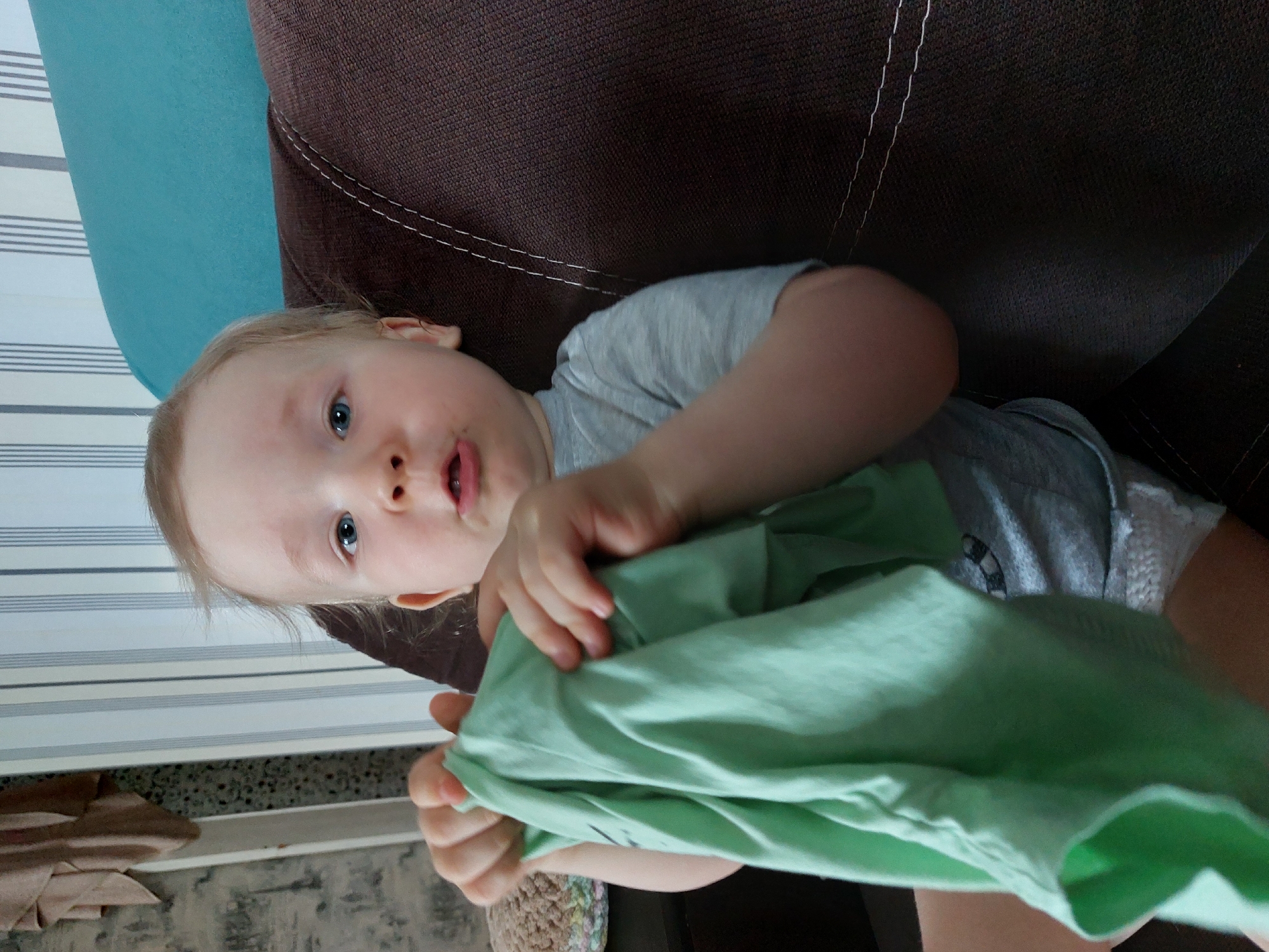 A young child, likely a toddler, with light blonde hair and blue eyes, is captured looking directly at the camera. The child is positioned on a brown sofa or armchair, which features visible white stitching and is partially covered by a turquoise cushion or fabric. The child's body is oriented diagonally across the frame.
The child wears a light grey t-shirt, which has a subtle, indistinct graphic in white and black on the chest. With both hands, the child actively grasps and holds a crumpled piece of light green fabric, possibly a garment. A small smudge is visible above the child's upper lip.
The background reveals a wall covered with striped wallpaper, featuring alternating light and dark horizontal bands. Below the wall, a section of the floor, which has a speckled or patterned surface, is visible. A folded light brown towel or blanket rests on the floor to the left, and a small, multicolored crocheted item is partially visible near the child's arm.
The setting appears to be an indoor domestic environment, possibly a living room or nursery, indicative of a casual moment at home. The lighting is soft and even, suggesting daytime. The child's expression is neutral and engaged with the camera. There is no visible text in the image. The location context provided is Chaikovsky, Russia.