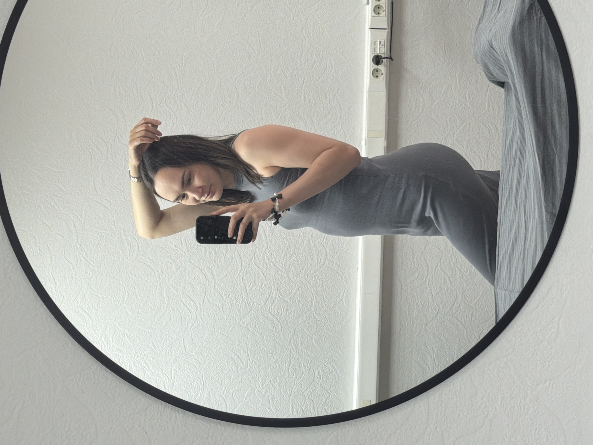 A young woman is reflected in a round mirror. She wears a gray dress and holds a smartphone in her left hand, appearing to take a selfie. Her right hand rests on her head. The background consists of a white wall, a white cable duct with electrical sockets, and a gray curtain partially visible in the mirror. The photograph was taken in Chaikovsky, Russia.