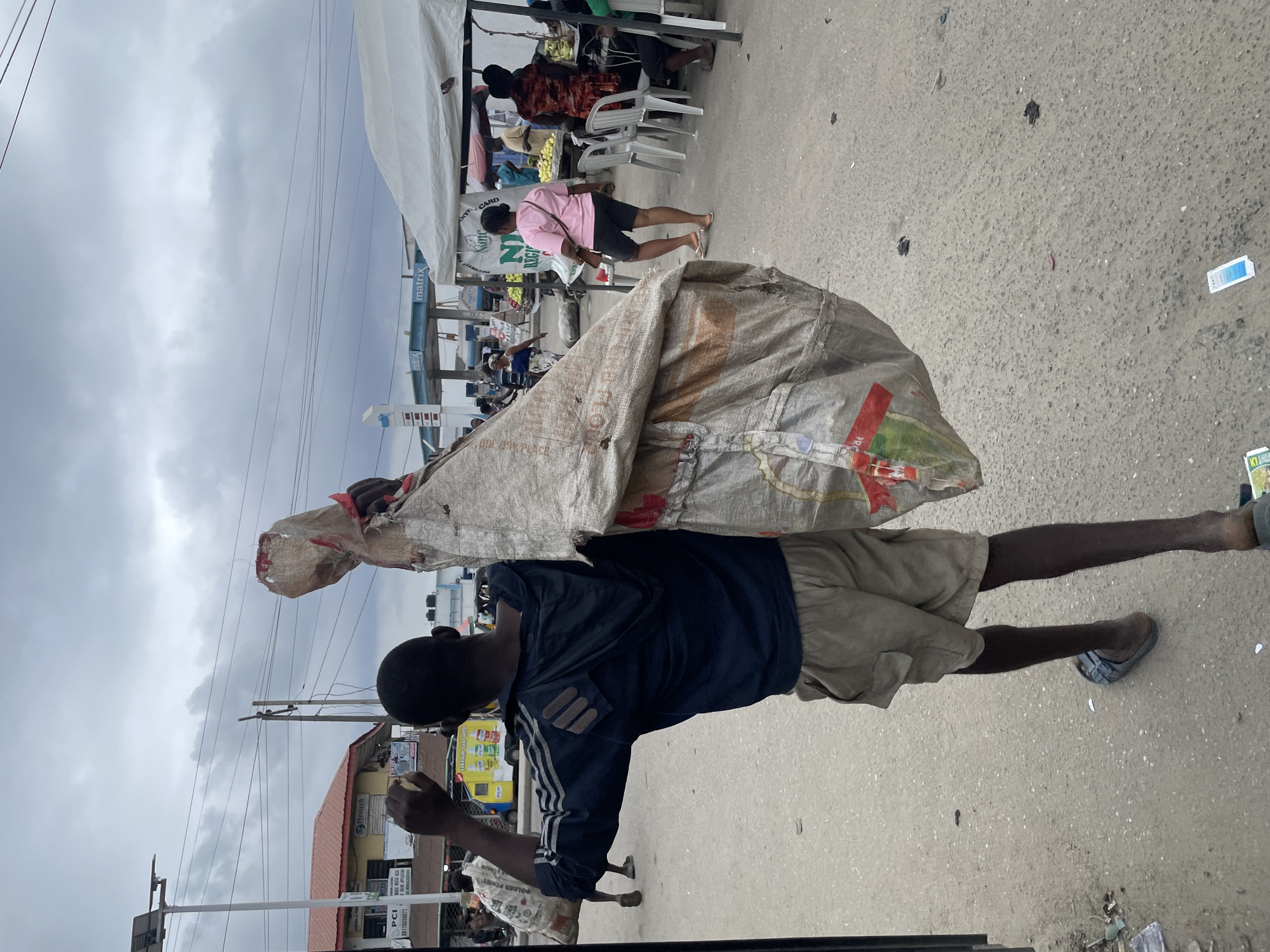 In Ado, Nigeria, on a cloudy day, a young person is captured from behind, carrying a large, worn, woven bag over their right shoulder. The bag appears to be filled with items, with a red tie visible at its opening. The individual is wearing a dark blue hooded jacket with white stripes on the sleeves and light brown cargo shorts. They are also wearing a pair of dark-colored sandals. The scene takes place on a paved road, which is somewhat littered. In the background, there is a bustling market area with white tents, people shopping and interacting. A gas station with the "Matrix" logo is visible in the distance, as are several buildings and streetlights. The overall atmosphere suggests a typical day in a Nigerian city's commercial district.