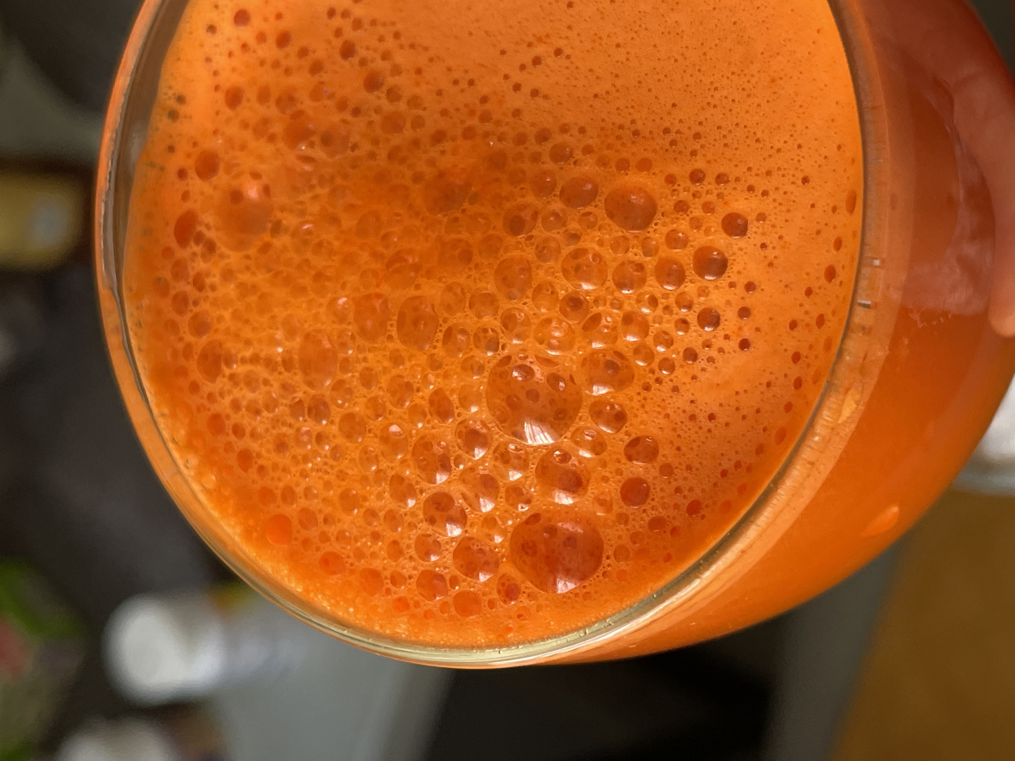 An overhead shot depicts a glass filled with a vibrant orange liquid, possibly juice. The liquid is thick and frothy, with many small bubbles on the surface. A hand holds the glass. The image is blurry and out of focus in the bottom right corner. The location and city of Sochi, Russia cannot be determined from the image.