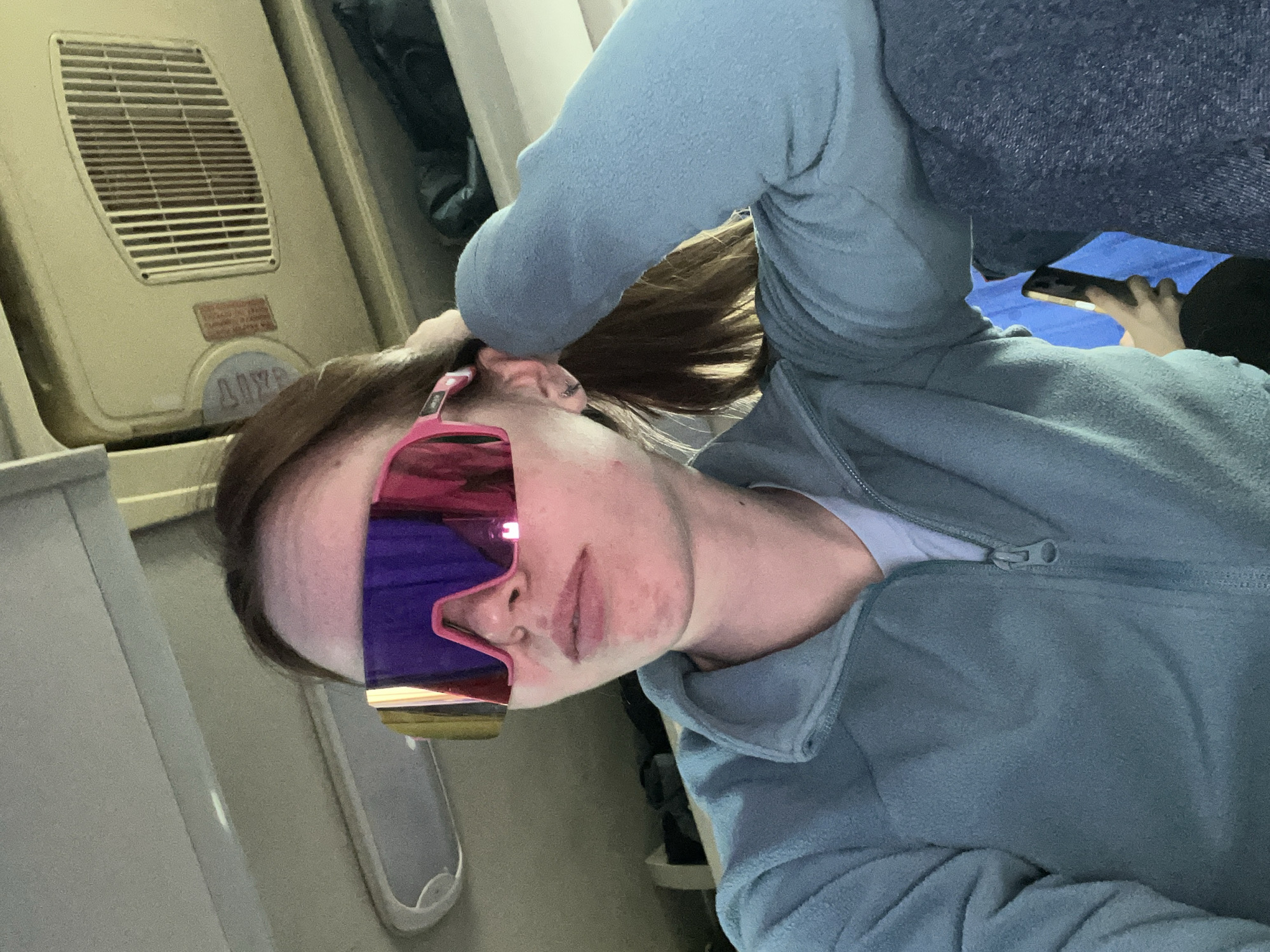 A woman with brown hair pulled into a ponytail is sitting on what appears to be an airplane. She is wearing large, mirrored sunglasses with a pink frame and a blue sweatshirt. She is holding the side of her head and looking at the camera. To her left is the interior wall of the aircraft, which includes a vent panel. Behind her to the right is another person, partially visible, looking at a phone. There are no indicators to confirm this image was taken in the city of Нижний Пальник, Russia.