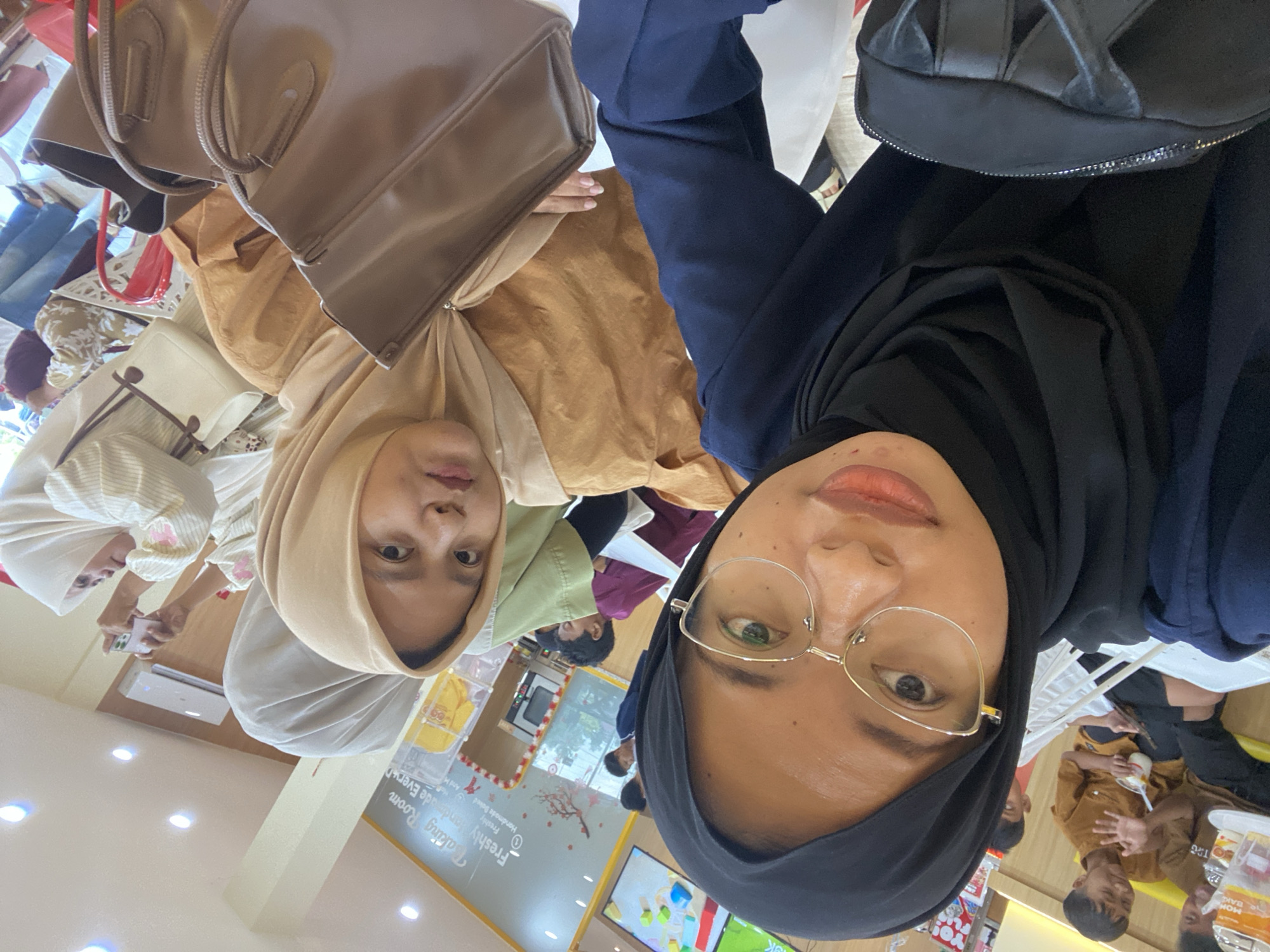 The image appears to be a selfie taken indoors, possibly at a cafe or bakery named "Baking Room". The foreground is dominated by two young women wearing hijabs. The woman on the left is wearing a black hijab and a dark blue top, with gold-rimmed glasses. She is holding a black bag. The woman on the right is wearing a light brown hijab and a tan top. She is holding a brown handbag. In the background, several children and adults are visible. Some children are sitting at a table, and one appears to be holding a cup. Another woman in the background is taking a photo with her phone. The environment suggests a casual dining or social setting with light-colored walls and artificial lighting. There are signs and decor visible on the walls, including one that says "Baking Room". Based on the context of "Kembang Harum, Indonesia", it is likely that this establishment is located in that area.