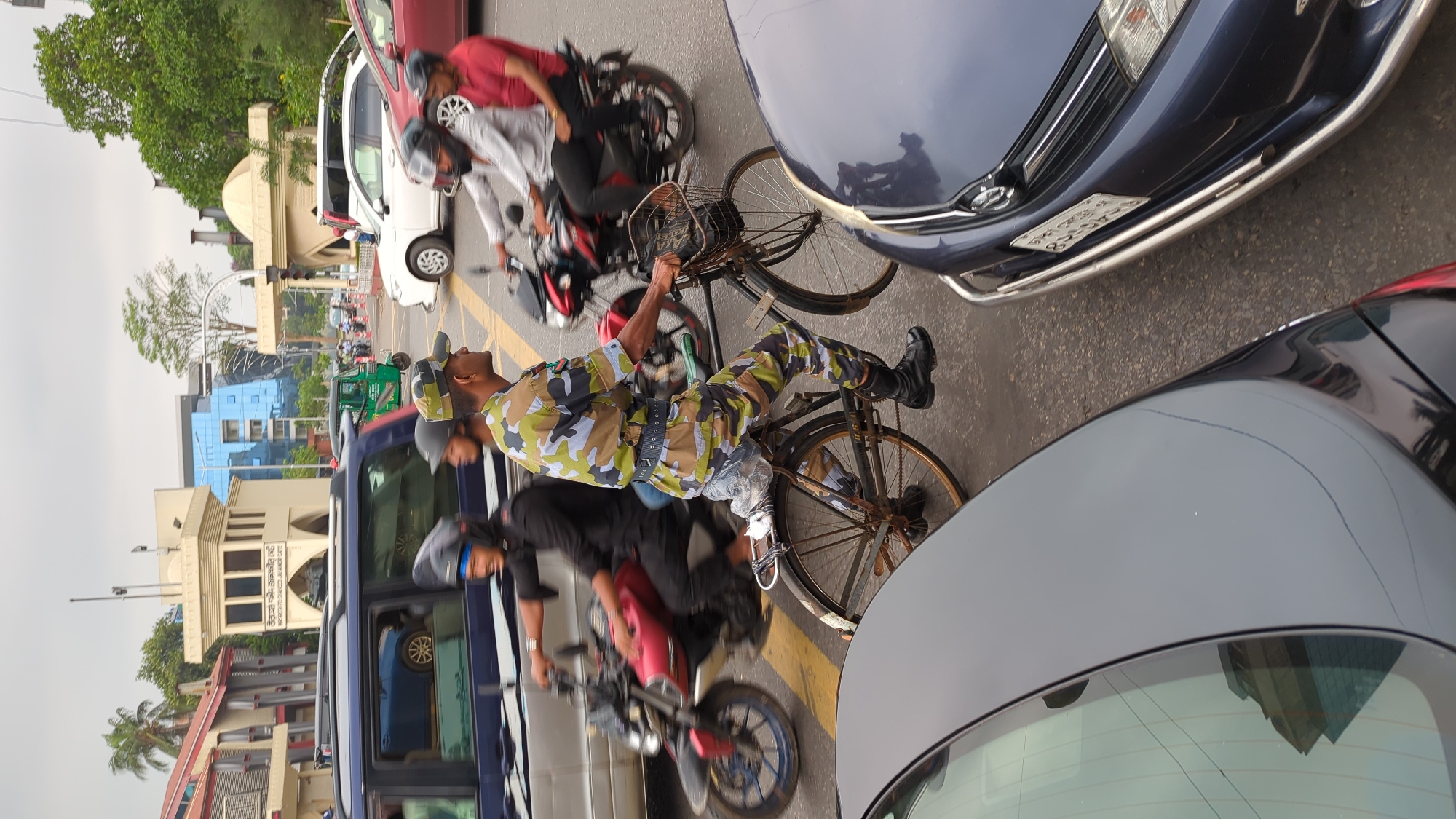 The image captures a busy street scene in Dhaka, Bangladesh. A traffic police officer in camouflage uniform and helmet rides a bicycle amidst heavy traffic. He is surrounded by cars, motorcycles, and a bus, all competing for space on the road. The officer looks upwards. Other people are riding the motorcycles. A building with architectural details, including a dome, is visible in the background along with modern glass buildings, indicating a commercial or administrative area. A license plate is visible on one of the cars in the scene, partially obscured. Yellow traffic lines mark lanes on the street. The overall impression is of a congested urban environment with active movement of vehicles and people.
