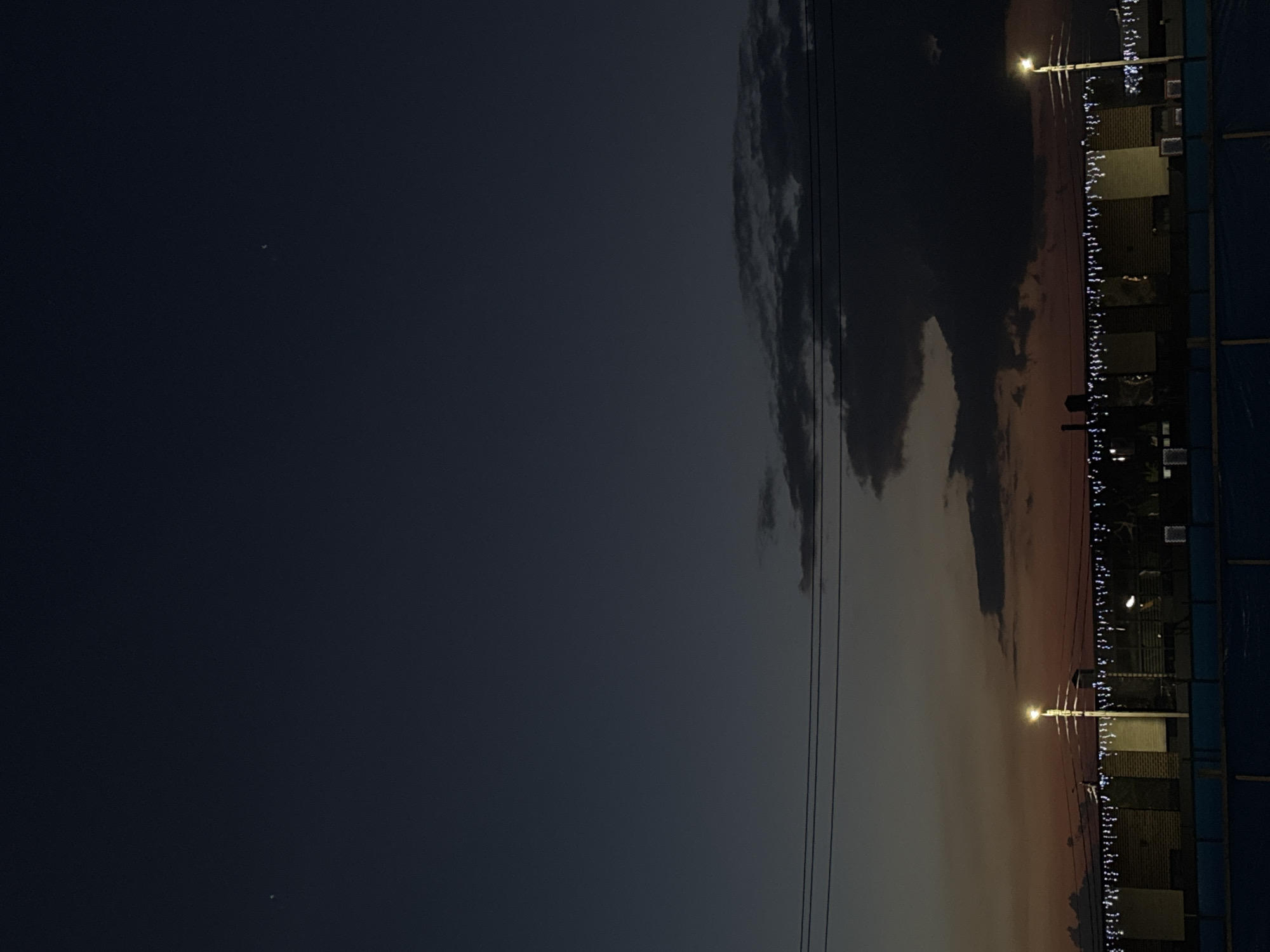 The image depicts a twilight scene in Mikhaylovsk, Russia. The sky is a deep blue, transitioning to hues of orange and red near the horizon, indicating the sunset or sunrise. Dark, textured clouds are visible in the middle ground, obscuring parts of the sky. Three power lines stretch horizontally across the frame. Below the power lines, a building or structure with a brick facade is illuminated by strings of white lights draped along its top. A tall lamppost with a single light source stands prominently in the lower right portion of the image. In the distance, other structures and possibly more lighting can be discerned. The foreground shows a blue barrier or fence. Two faint, bright points of light are visible in the upper portion of the sky, likely stars or planets. No people or animals are visible in the image.