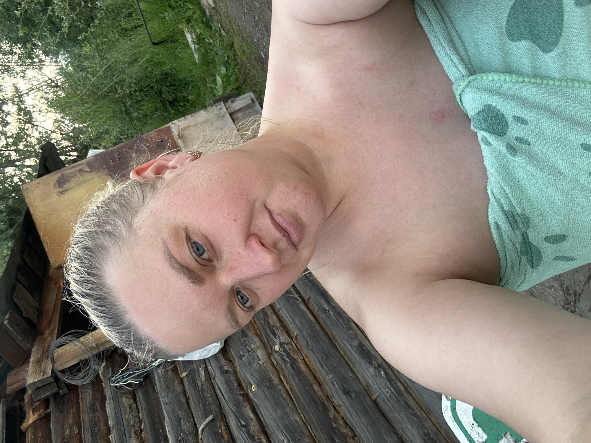 A woman is pictured from the chest up, outdoors, in front of a rustic wooden structure. She is wrapped in a light green towel adorned with a dark green polka dot pattern. Her blonde hair is pulled back from her face, and she wears a small, gold-colored earring in her left ear. The background includes the wooden structure and lush green foliage. The setting appears to be rural or in a village, consistent with the outskirts of Chaikovsky, Russia.