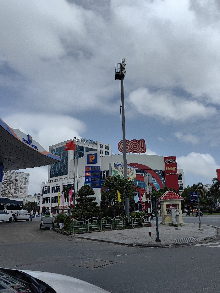 The image depicts a bustling urban street scene in Thuận Hóa Ward, Vietnam, under a partly cloudy sky. In the foreground on the left, a "Petrolimex" gas station canopy is visible, with two white vehicles and a person in a light blue shirt near the fuel pumps. Adjacent to the gas station is a multi-story building featuring blue-tinted windows, a "P" logo, and a Vietnamese flag flying on a pole. An electronic display board shows fuel prices. A small, white utility cart is parked on the curb nearby.
In the midground, a large commercial building, identified by a prominent red "GO!" logo at its peak, dominates the right side of the frame. Below the main logo, "Kohn" and "LOTTERIA" signs are visible. This building has a modern, multi-colored facade. A tall, grey utility pole with an array of fixtures at its summit stands between the gas station and the "GO!" building.
At a street corner in the center-right, a small, red-roofed kiosk or booth is situated on a paved island, next to a red and white striped traffic light pole. The area also features a manicured green space with trimmed hedges and small trees, bordered by a green metal fence. The street is paved, and multiple motorcycles, some with riders, are visible on the road, indicating active traffic. Palm trees and other buildings are present in the background.