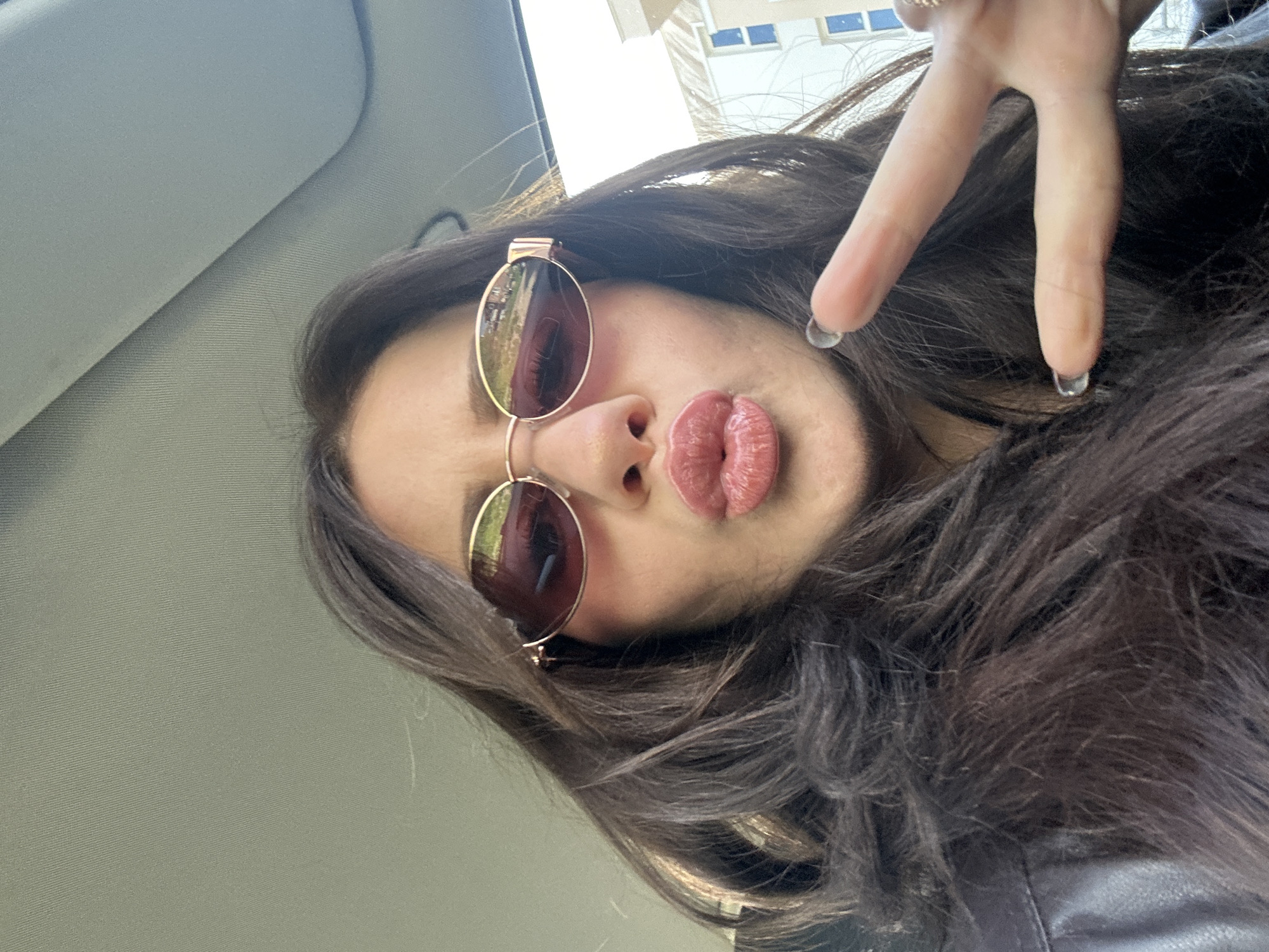A close-up selfie captures a person, likely female, inside a vehicle during daytime. The person has long, dark, wavy hair and is looking upwards at the camera, holding it from a low angle. They are wearing round, reflective sunglasses with reddish-brown lenses and gold frames, resting on their nose. Their lips are puckered in a "duck face" pose. The right hand is raised, making a peace sign (V-sign) with two fingers, and clear or transparent acrylic nail tips are visible on the fingernails.
The background shows the light grey fabric of a car's headliner and sun visor on the left and center. To the upper right, through a window, a distant building with rectangular windows is partially visible against a bright sky, suggesting an urban or suburban environment. A dark, possibly leather, garment (like a jacket) is visible on the person's right shoulder and arm. The lighting is bright, indicating it is daytime. The overall impression is casual and playful, taken within a car in Plovdiv, Bulgaria. There is no visible text in the image.