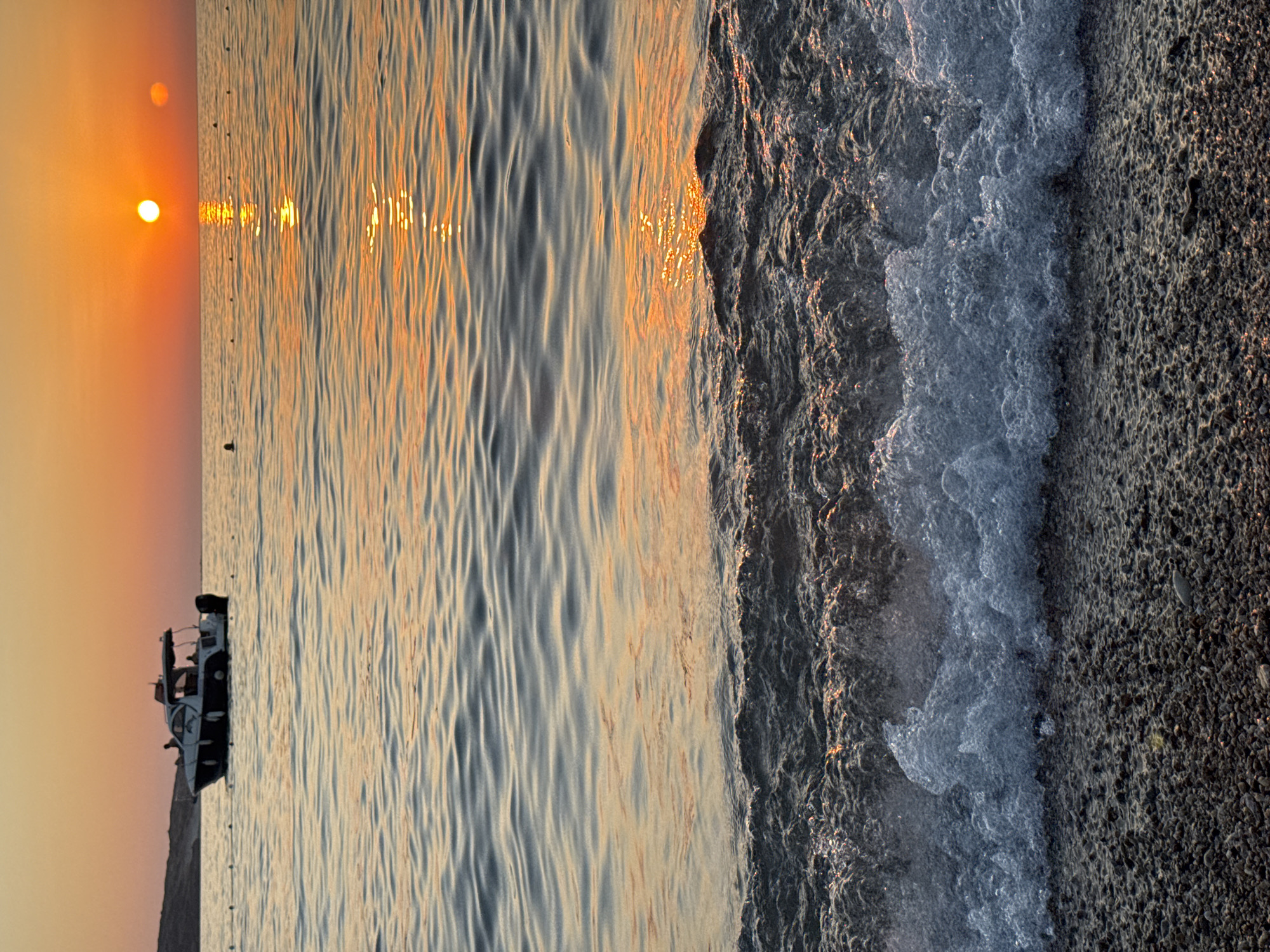 A medium shot captures a tranquil scene at dusk in Foça, Turkey. The sun, a bright orange orb, hovers just above the horizon, casting a warm glow across the water and the sky. Its reflection shimmers on the gentle waves, creating a golden pathway. In the middle ground, a white motorboat with a dark hull is anchored, with a few figures visible on board. The boat appears to be a day cruiser or yacht, equipped with an awning over the cockpit. The water is calm, with small ripples extending to the shore, where a gravel beach is visible in the foreground. The overall impression is one of peaceful serenity as day transitions into night.