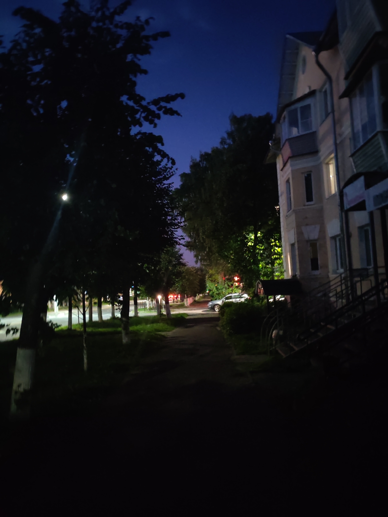 A nighttime photograph captures a street scene with a building and trees. The sky is a deep blue, suggesting twilight or early night. On the right, a multi-story building with light-colored walls and balconies is visible. Its windows are illuminated, indicating occupancy. A dark staircase leads up to an entrance on the right side of the building.
To the left of the building, a paved walkway runs alongside a row of trees that cast dark shadows. The trees are silhouetted against the sky, and some have white painted bases, common in Russian cities. Beyond the trees, streetlights and traffic lights are visible, casting a faint glow on the road. A silver car is parked on the street. The overall impression is of a quiet, residential street at dusk in a city.
This scene is consistent with a street in Vladimir, Russia, given the architectural style of the building, the presence of street lighting, and the general urban landscape. The photograph appears to have been taken from a slightly elevated position, looking down the street.