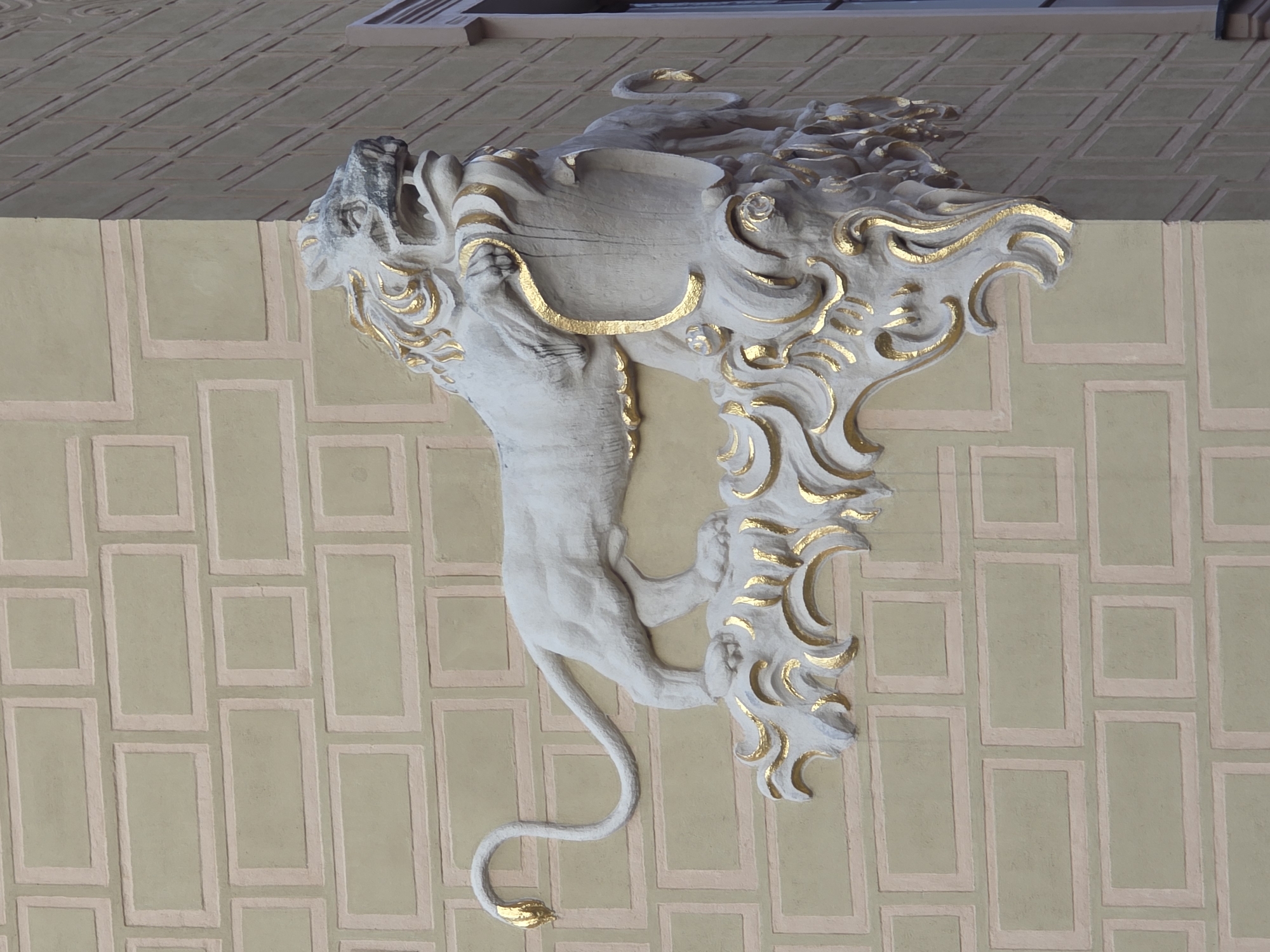 A sculpted white lion with gold accents is attached to the corner of a building with a pale yellow facade decorated with a brick pattern. The lion is depicted in profile, with its mouth open and its front paws resting on an ornate, gold-trimmed base. Its tail curves upwards and its ears are pointed. The building's facade is painted to resemble light brown bricks with pink mortar lines. To the right of the lion, the building has a different facade with diagonal lines, suggesting a change in architectural style or material. The scene appears to be an outdoor architectural detail in a city.
