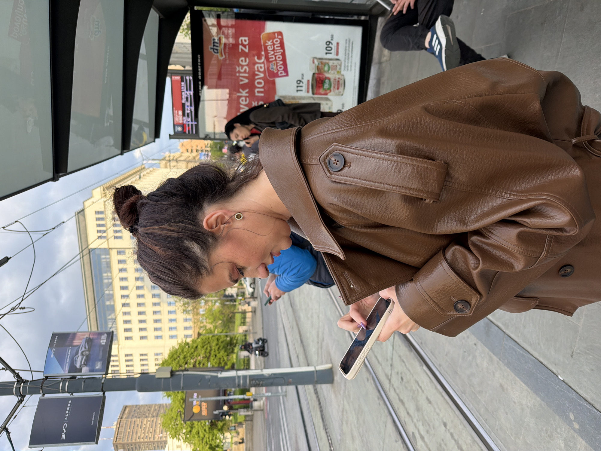 This media file presents a daytime urban street scene in Belgrade, Serbia, under a partly cloudy sky. The central figure is a young woman, seen in profile, intently focused on her smartphone. She wears a brown faux leather jacket, featuring visible buttons on the collar and cuffs, and her dark hair is neatly tied back in a bun. Her attention is directed downwards towards her device, suggesting she is checking messages or browsing.
The setting reveals a busy city environment with prominent tram tracks running through the foreground and midground. In the background, several multi-story city buildings are visible, along with lush green trees. A street pole adorned with advertisements, including one for "BYD AL PROS," stands in the midground. To the right, a bus stop shelter with glass panels features a large billboard. Another individual, partially obscured and wearing a blue top, is also seen looking at a smartphone behind the main subject. Further back, the lower half of another person, wearing dark pants and sneakers with blue stripes, is visible.
Visible Serbian text on the billboard includes "dm," "uvek više za novac" (always more for money), and "uvek povoljno,-" (always affordable), accompanied by prices "119" and "109" for product cans, one labeled "Bio." Reflections on the glass panels of the bus stop show partially reversed text like "Болорис" and "GUATTRO." The scene captures a typical moment of urban daily life, with people navigating and interacting within the city landscape.