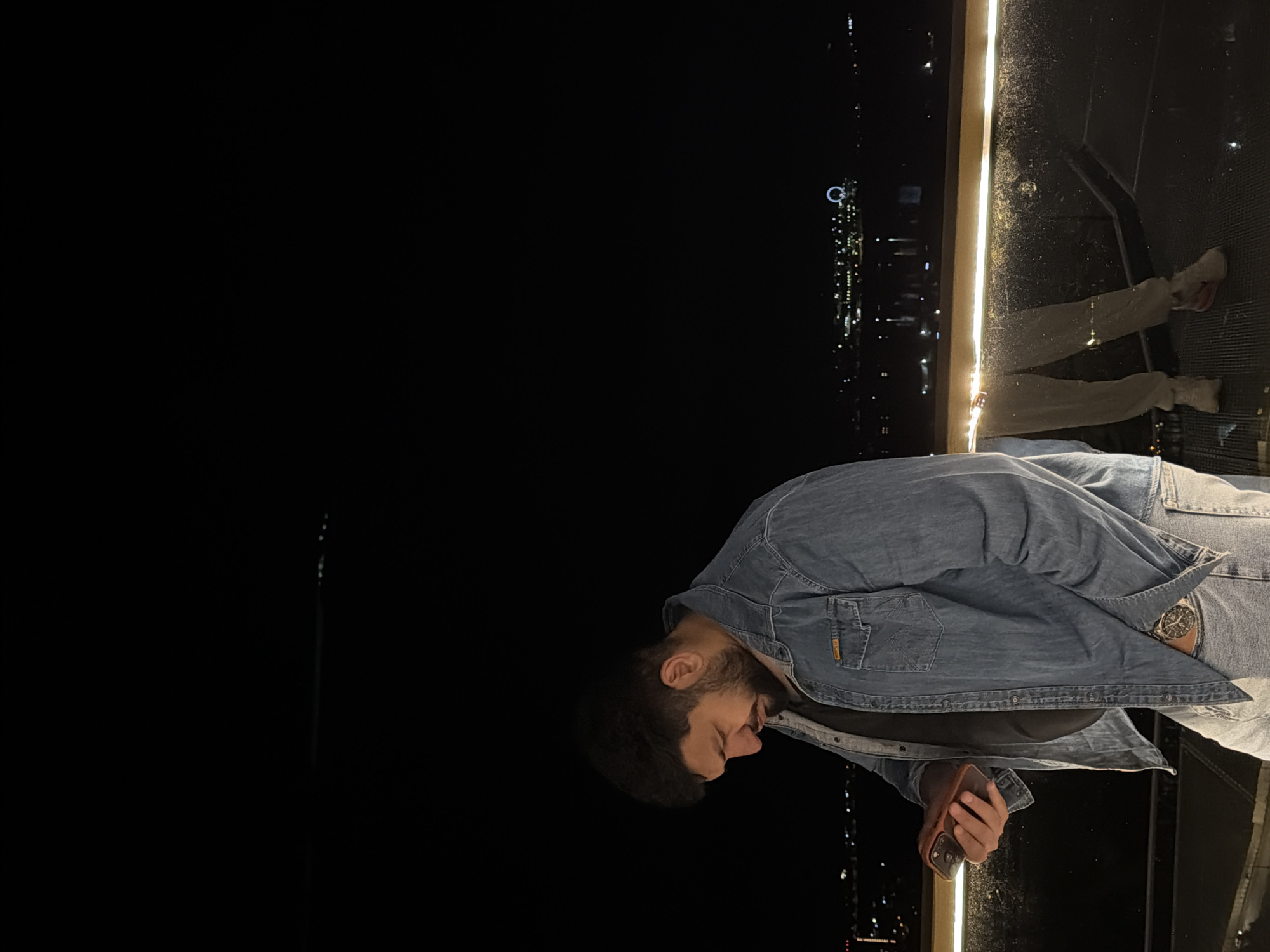 A man with a beard is looking down at his phone at night. He is wearing a denim jacket, and a watch is visible on his left wrist. He is standing on an observation deck with his back toward the city of Krasnoyarsk, Russia, which is visible in the background. The city lights twinkle in the distance. The floor of the observation deck is a dark material. There is a light or railing adjacent to the edge of the observation deck. The man’s legs and feet are visible, indicating he is wearing casual pants and shoes. The majority of the image background is a dark night sky.