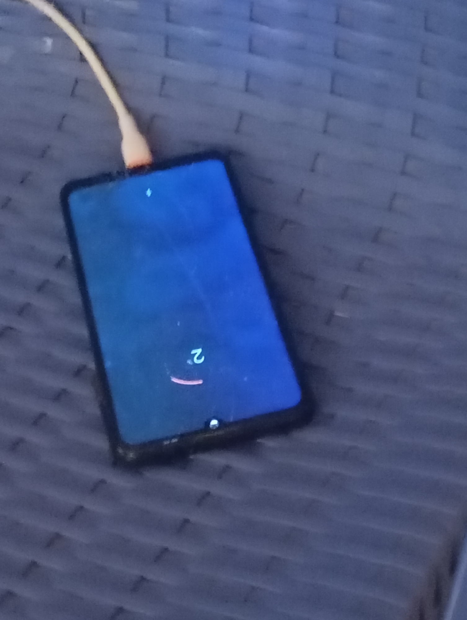 A smartphone is charging on a dark blue, textured surface. The phone's screen displays a bright blue background with a small white icon at the top and a large white number "2" with a red curved line beneath it. A light-colored charging cable is plugged into the phone. The background surface appears to be an outdoor furniture set or a woven mat. The location is Unknown, Unknown.