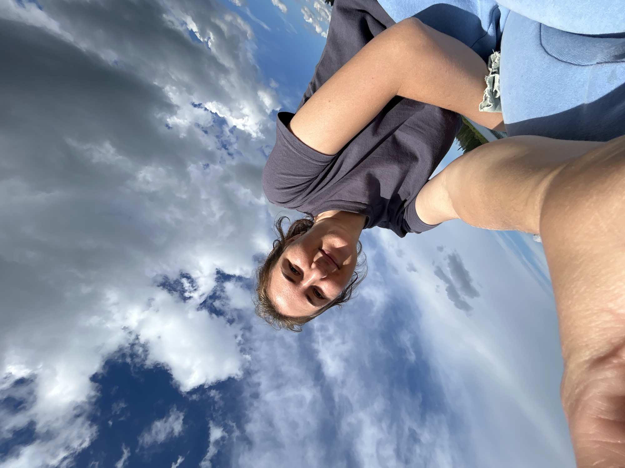 The media file is a selfie taken from a low angle, looking up at the sky. The main subject is a person, likely female, with brown hair, who is smiling. They are wearing a dark grey t-shirt and light blue shorts with a frayed hem. The background is dominated by a dramatic sky with a mix of dark, stormy clouds and bright blue sky with scattered white clouds. The lighting suggests it is daytime, possibly late afternoon or early evening, given the dynamic cloud formations. The perspective creates a sense of looking up from a ground-level position, possibly while lying down or sitting on the ground. The overall mood is casual and relaxed, with the subject appearing happy and enjoying the outdoors. The location context provided is Верх-Рождество, Russia. No visible text is present in the image.