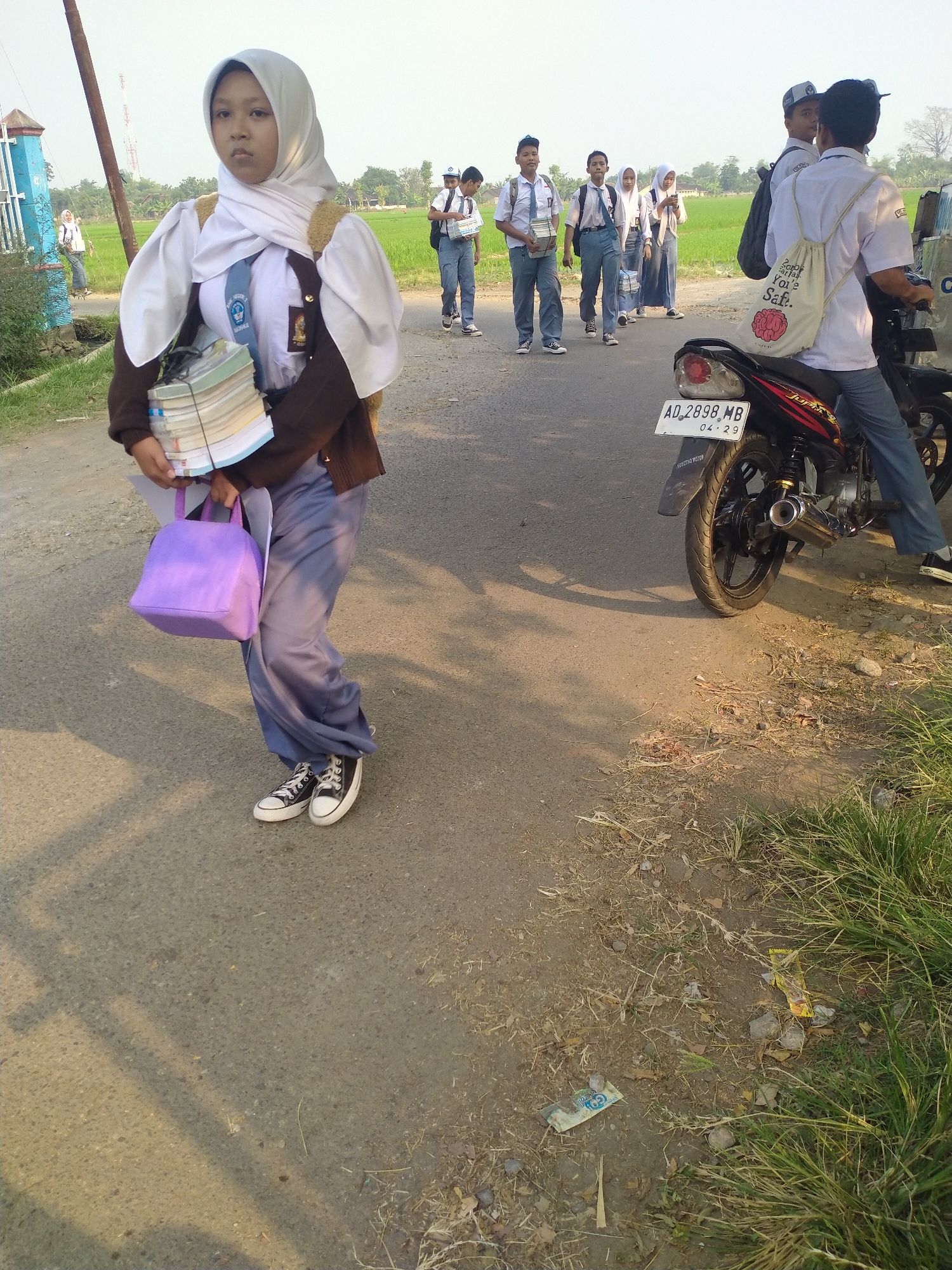 In the village of Begajah, Indonesia, a young female student wearing a hijab and a school uniform, consisting of a white shirt, brown blazer, and grey pants, stands on a paved road. She carries a stack of books in her left arm and a purple handbag in her hand. Several other students in similar uniforms are walking on the road in the background, some carrying books. A motorcycle is parked on the right side of the road. The environment appears to be rural, with a rice field visible in the background. The sunlight casts shadows on the road, indicating it is either morning or late afternoon.