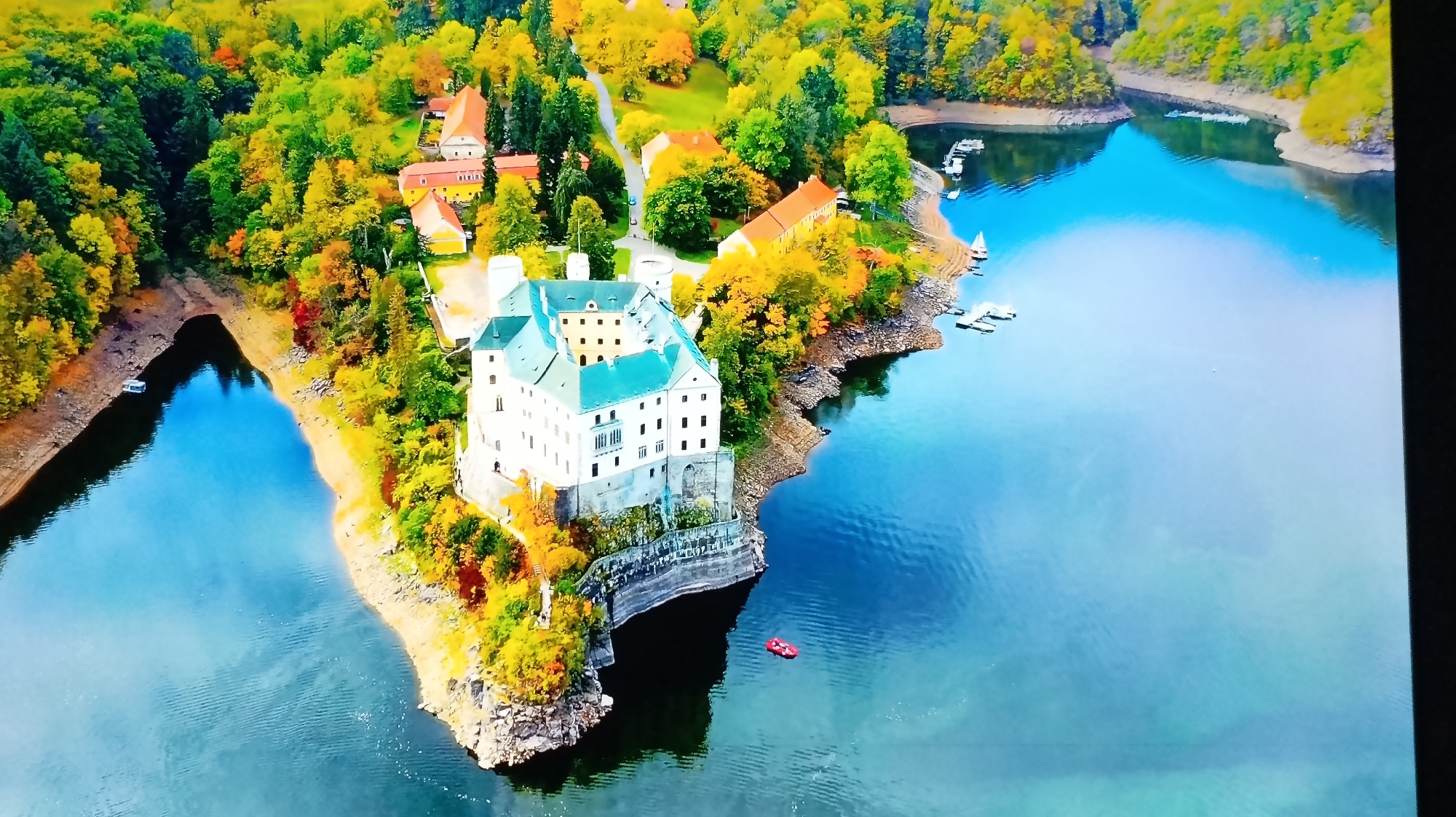 An aerial drone shot captures a picturesque autumn landscape with a white castle perched on a rocky peninsula extending into a serene blue lake. The castle features a green tiled roof and multiple windows. Surrounding the castle and along the lake's edge are trees displaying vibrant fall foliage in shades of yellow, orange, and red. Several boats are visible in the lake, including small sailboats and what appears to be a red paddle boat near the castle. In the background, additional buildings with orange roofs are nestled among the trees, suggesting a small village or estate. The water is calm, reflecting the colorful trees and sky. The scene is located in the city of Roghi, Moldova.