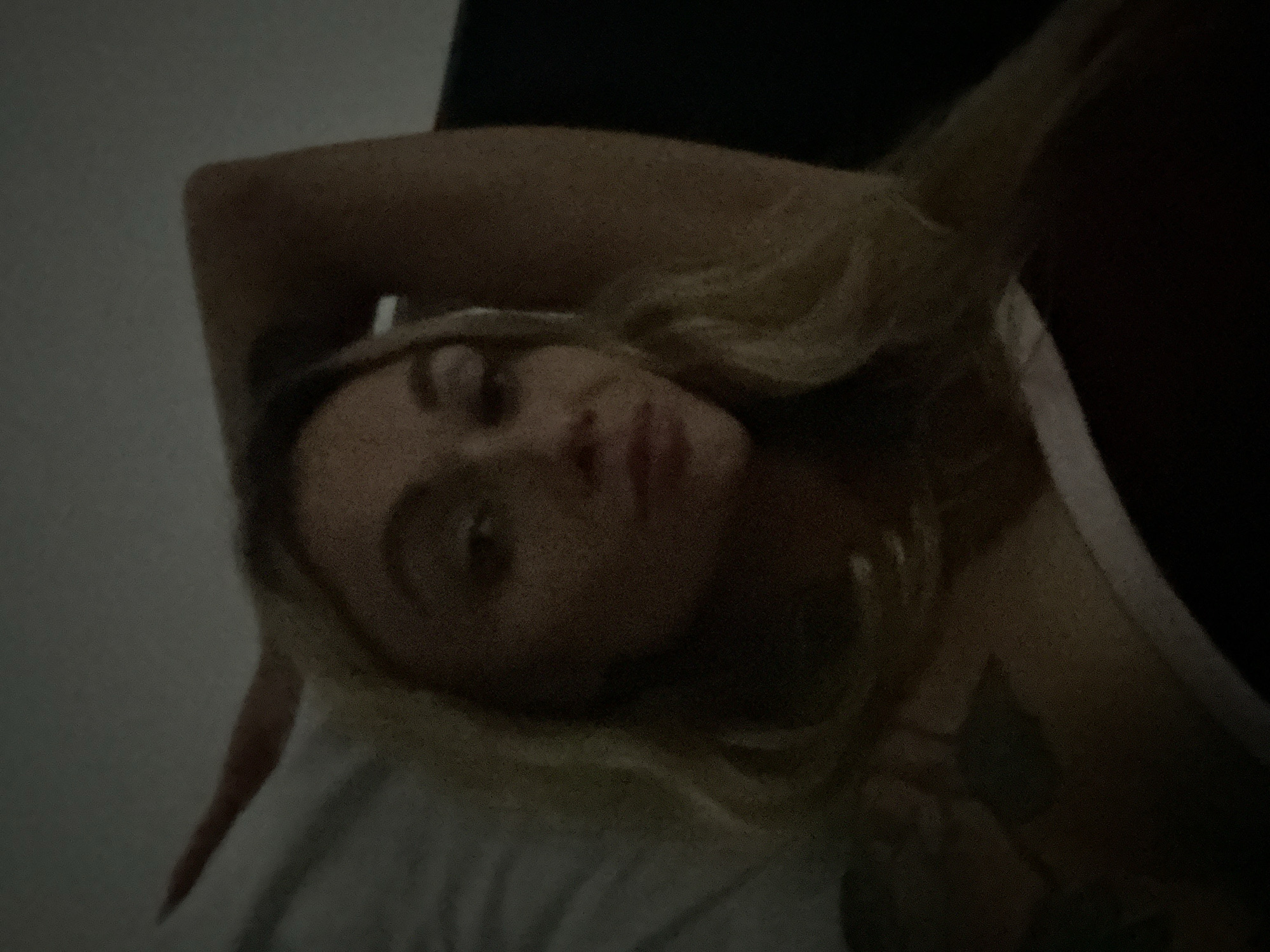A low-light, close-up image captures a person, likely a woman with blonde hair, lying down indoors. Her head is slightly tilted, and she looks towards the camera with a neutral to contemplative expression. Her eyes are open, and her lips are softly closed.
Her left arm is bent, with her hand resting near her head, while her right arm and shoulder are partially visible on the right side of the frame. A white or light-colored strap, possibly from clothing like a tank top, is visible over her right shoulder. A tattoo with a dark, organic or floral design is visible on her right arm/shoulder area.
She is resting on a white or light-colored pillow and bedding, indicating a bedroom setting. The lighting is very dim, resulting in a dark background and a generally grainy image, suggesting it was taken at night or in a poorly lit room. No specific location cues are visible within the image, but the context provided places it in Đồng Nai, Vietnam. There is no visible text.