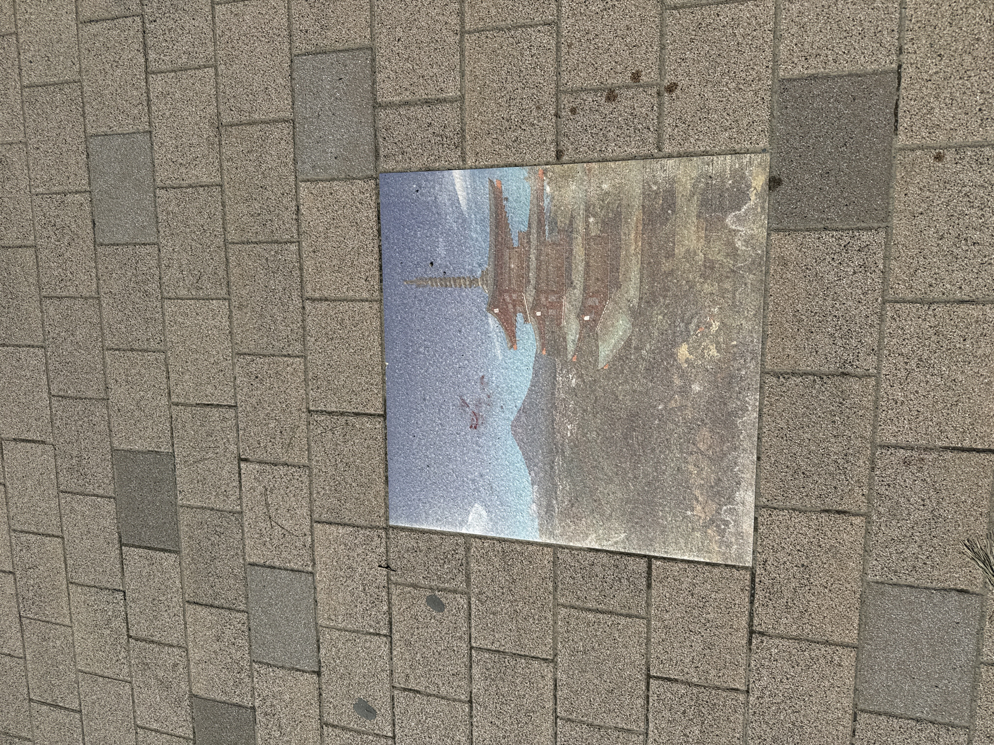 The image provides an overhead view of a paved ground surface, featuring a rectangular plaque embedded within it. The pavement is composed of numerous rectangular, light greyish-brown blocks with a rough, speckled texture, arranged in a staggered, brick-like pattern. Some blocks appear darker grey, possibly due to varying material or wear.
A rectangular plaque, positioned towards the center-right of the frame and slightly rotated, is the focal point. This plaque displays a faded, worn, and somewhat abstract image. The image on the plaque appears to be an inverted or reflected depiction of a traditional multi-tiered Japanese pagoda-style building, characterized by its dark brown or reddish-brown structure and distinct, tiered roofs against a lighter blue background, possibly representing a sky or water reflection. Subtle outlines that could be mountains are also visible in the background of the plaque's image. The plaque itself shows signs of weathering and a textured surface.
No people or direct activities are visible in the static scene. The setting is an outdoor paved area, likely a pathway or public space, illuminated by bright daylight. The ground appears dry. Given the location context of Fujiyoshida, Japan, the building depicted on the plaque is likely a local landmark, possibly shown as a reflection or an artistic representation. No visible text is present on the plaque or the surrounding pavement.