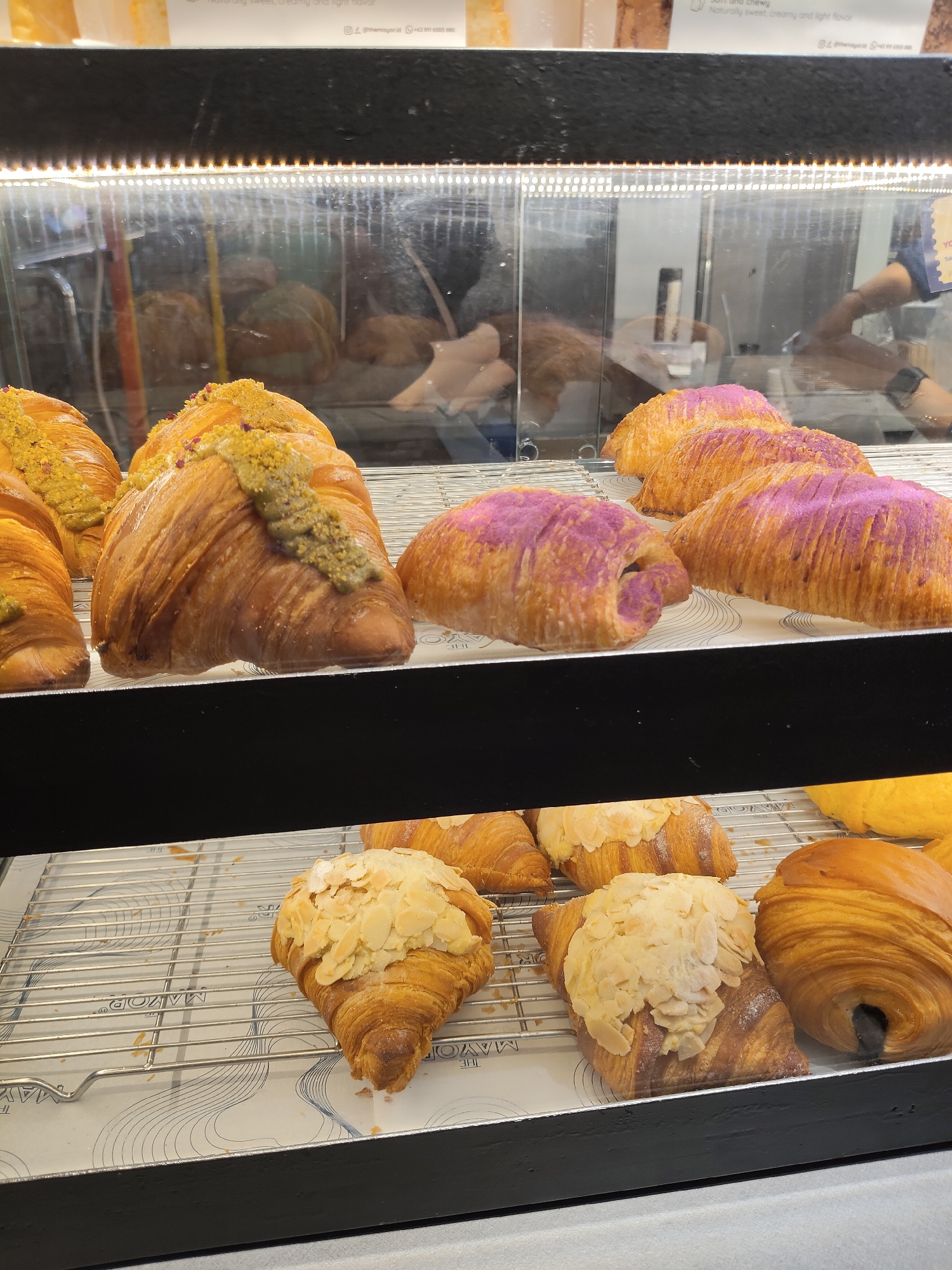 The image shows a display case filled with various types of croissants at a bakery. In the upper part of the display, several croissants are visible. One prominent croissant is topped with a green paste and what appears to be crushed pistachios. Next to it are croissants with a purple topping, possibly taro. In the lower section of the display, there are more croissants. Some are sprinkled with sliced almonds, while others are a golden brown color. A person's hand and arm, wearing a dark shirt, are partially visible in the background, suggesting a staff member is present. The display case is illuminated by overhead lights. The overall environment appears to be a retail setting for baked goods. The presence of the text "THE MAYOR" on the paper liner under the croissants suggests the name of the establishment. Based on the context provided by the prompt that the location is the City of Medan, Indonesia, this is a bakery in Medan.