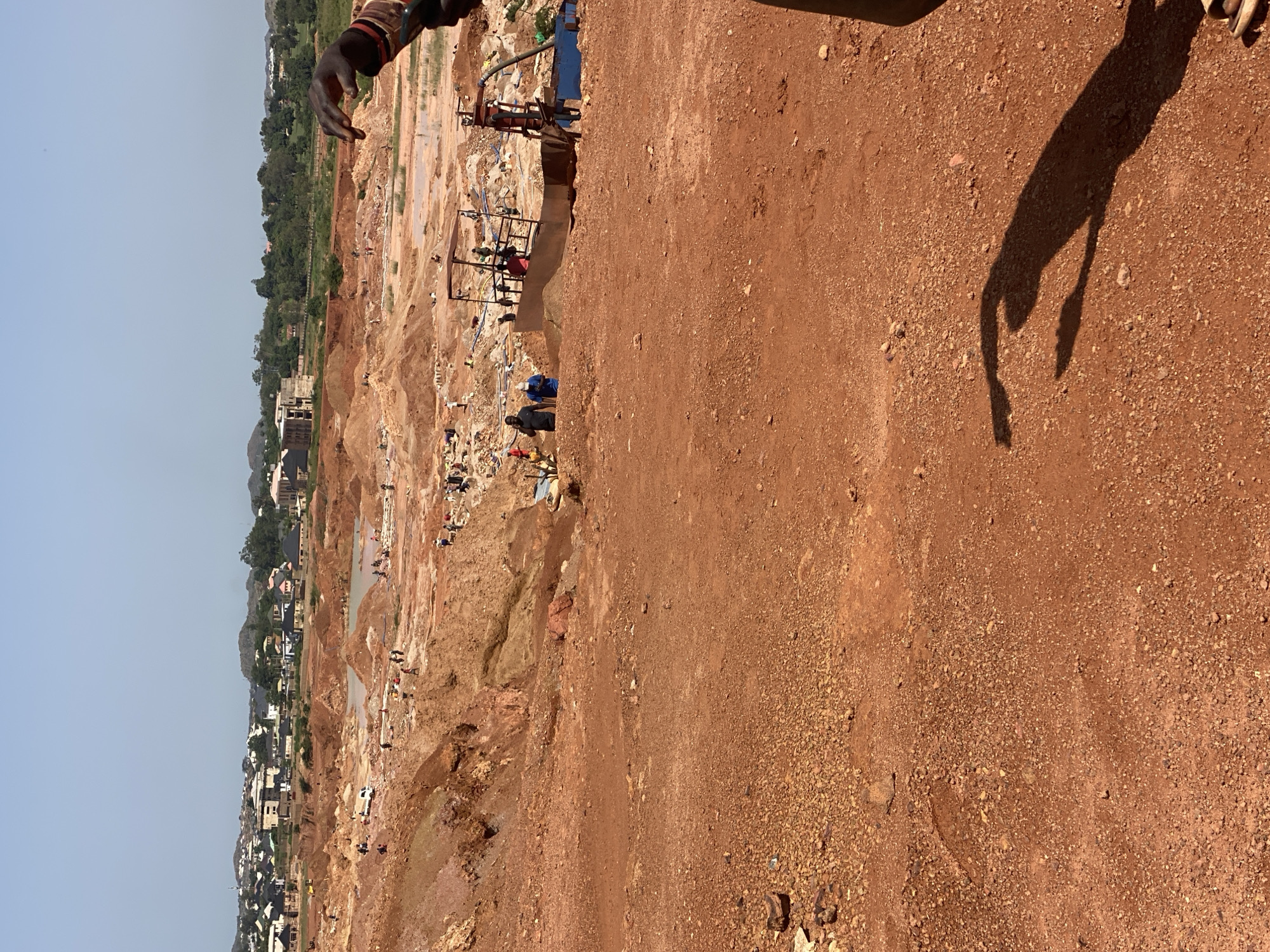 This image depicts an active mining site located in the city of Jos, Nigeria. The foreground is dominated by reddish-brown soil and small stones. In the mid-ground, the landscape transitions to a series of excavated pits and mounds of earth, indicating ongoing mining operations. Several people are visible scattered throughout this area, engaged in various activities. Some appear to be working in or around the pits, while others are near a piece of mining equipment. A white truck is parked in the mid-ground. In the background, the scene opens up to a more developed area with buildings, trees, and hills, suggesting the proximity of a residential or urban zone to the mining site. The sky is clear and blue. A person's hand and arm, adorned with bracelets, are partially visible in the upper right corner of the frame, along with a shadow cast by a person in the foreground.