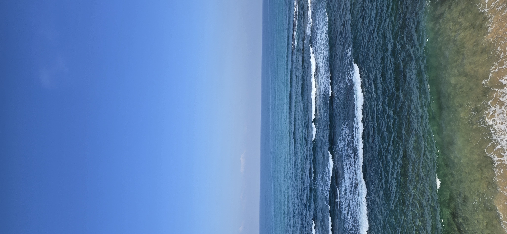 The image shows a view of the ocean under a clear blue sky. The water transitions from a deep blue in the distance to a lighter, greenish-blue closer to the shore, where waves are breaking. The shore itself is sandy and appears shallow, with foamy white water from the waves washing over it. There are no people or man-made objects visible in the scene.